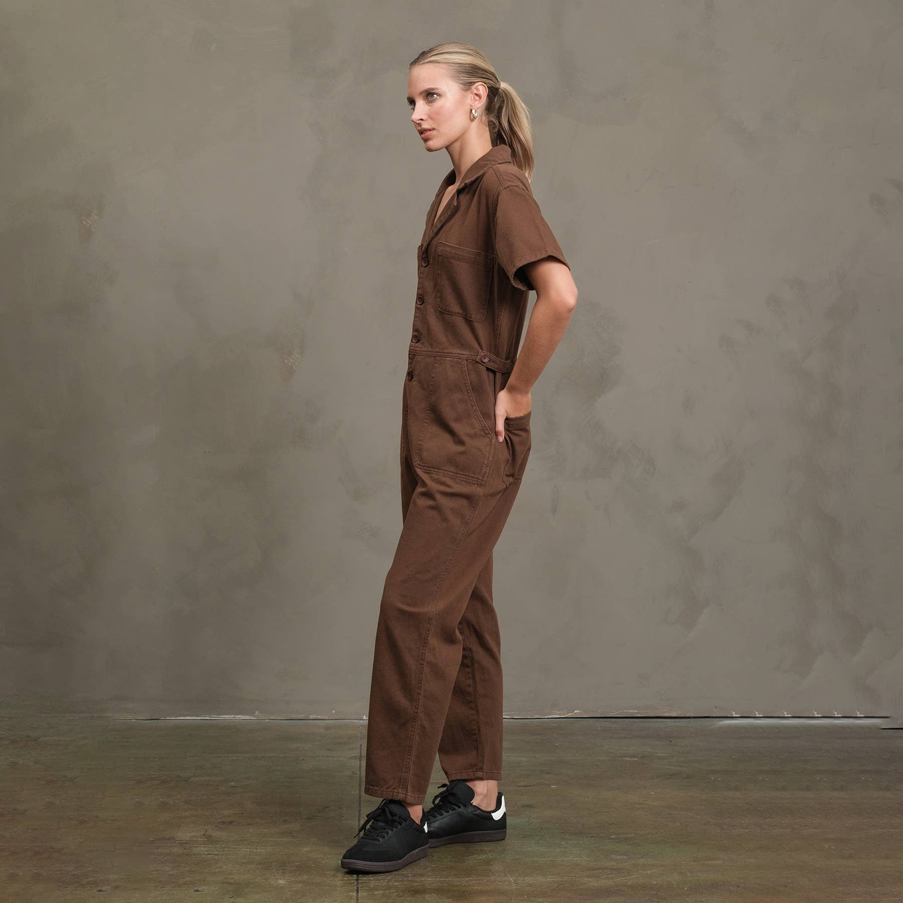 A woman stands in profile against a gray textured wall, wearing the RAISONNEL Field Jumpsuit (RJP1001) and black sneakers, with her hair pulled back and hands in her pockets.
