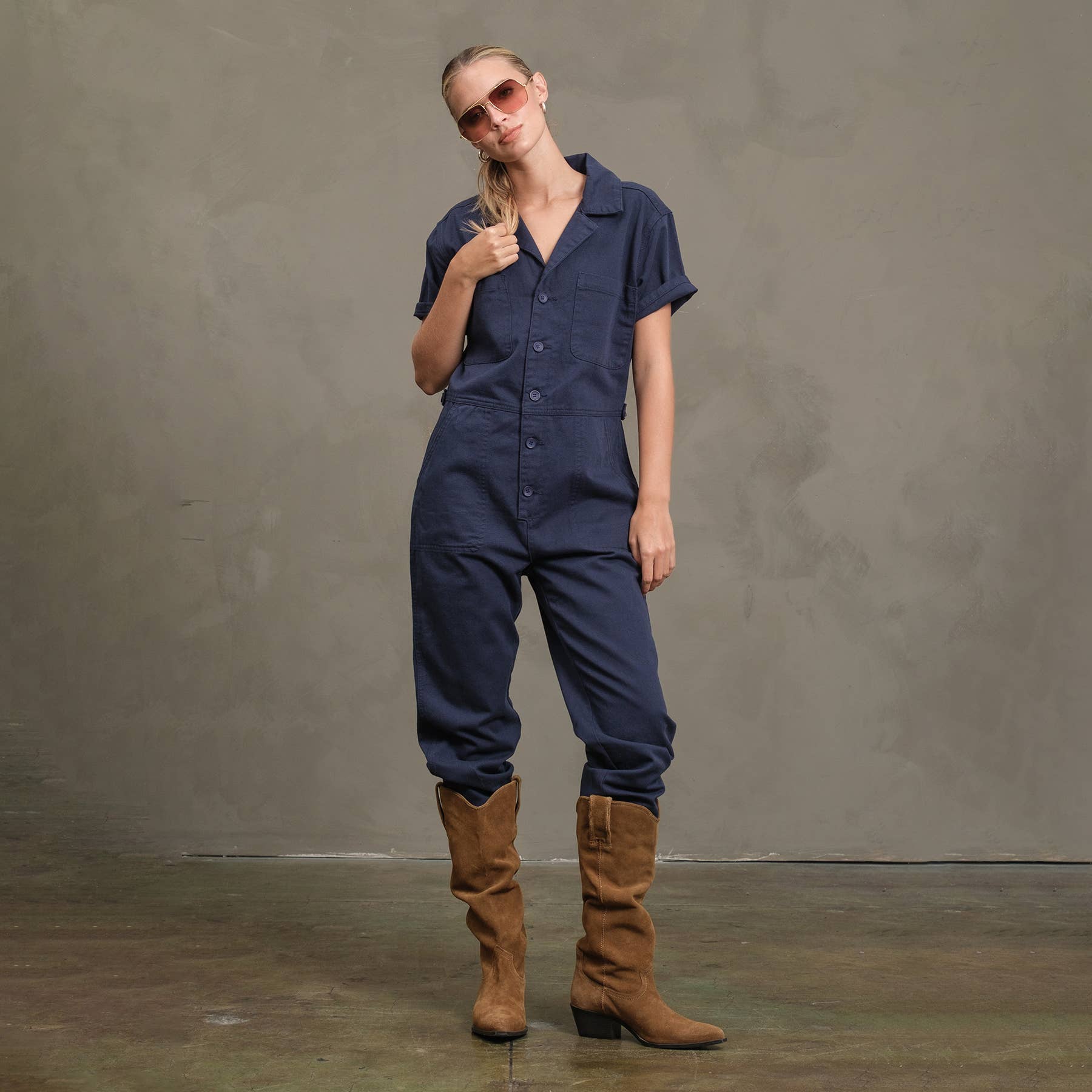 A person gives a thumbs up with a slight head tilt while wearing RAISONNEL’s Field Jumpsuit - RJP1001, sunglasses, and brown knee-high boots, standing against a plain gray background.