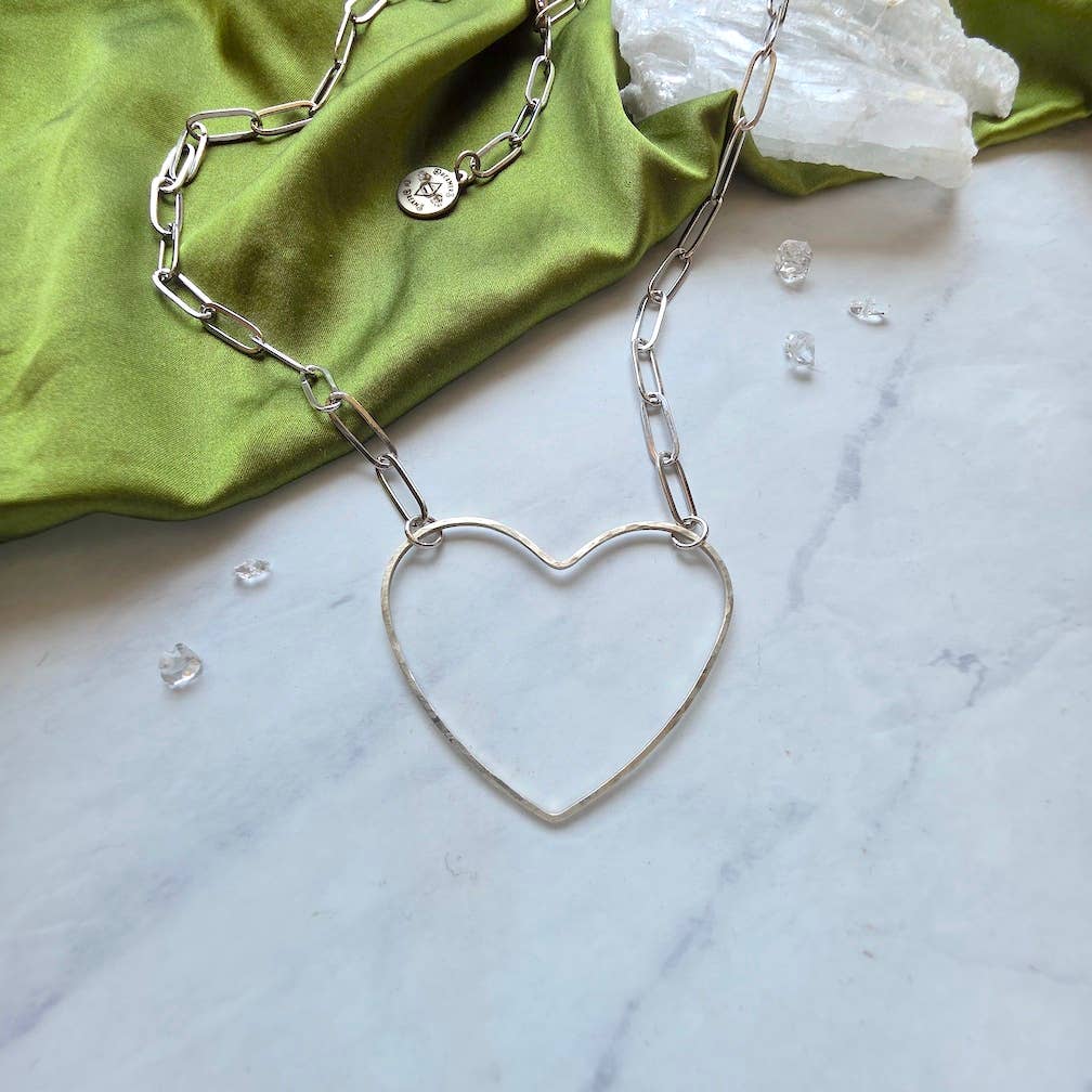 The Big Silver Heart -Paperclip Chain Necklace by Dreamers of Dreams features a large hammered heart charm on a paperclip chain, crafted from silver-plated brass and displayed on marble with green satin and clear crystals.