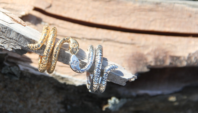The Rattlesnake Lotus Ring duo by bahgsu jewels—one in handcrafted gold brass, one in silver—coil artfully together atop weathered wood with a sunlit rustic backdrop, creating a striking minimalist statement.