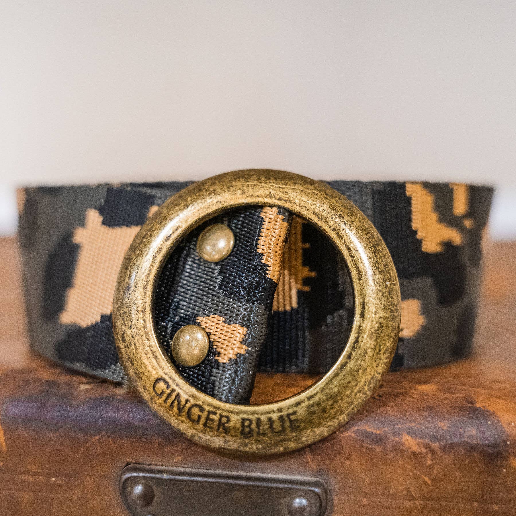 A close-up of the Wild Child Leopard belt by Ginger Blue highlights its leopard print fabric and large, round, antique brass cinch-style buckle engraved with GINGER BLUE, beautifully displayed on a wooden surface.