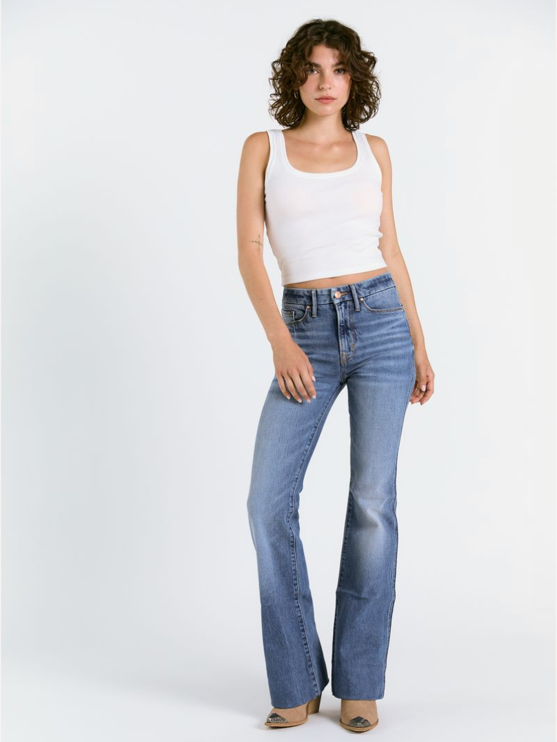 A woman with curly brown hair wears a white tank top, beige pointed-toe shoes, and Porter Blue Apparels WANDERER 5 POCKET FLARE IN MABEL WASH jeans, standing against a plain white background.