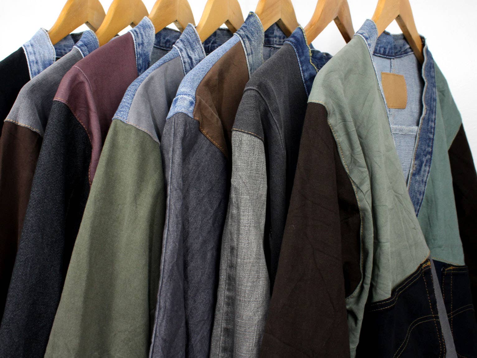 Row of Upcycled Patchwork Denim Jackets in various colors and mixed fabric textures hanging on wooden hangers.