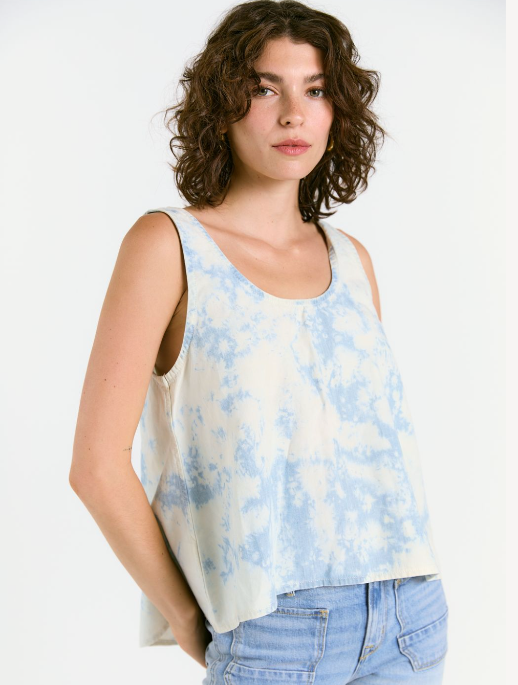 A woman with curly brown hair wears the eco-friendly WABI SABI TANK IN SKY TIE DYE by Porter Blue Apparel, paired with light blue jeans, standing against a plain white background.