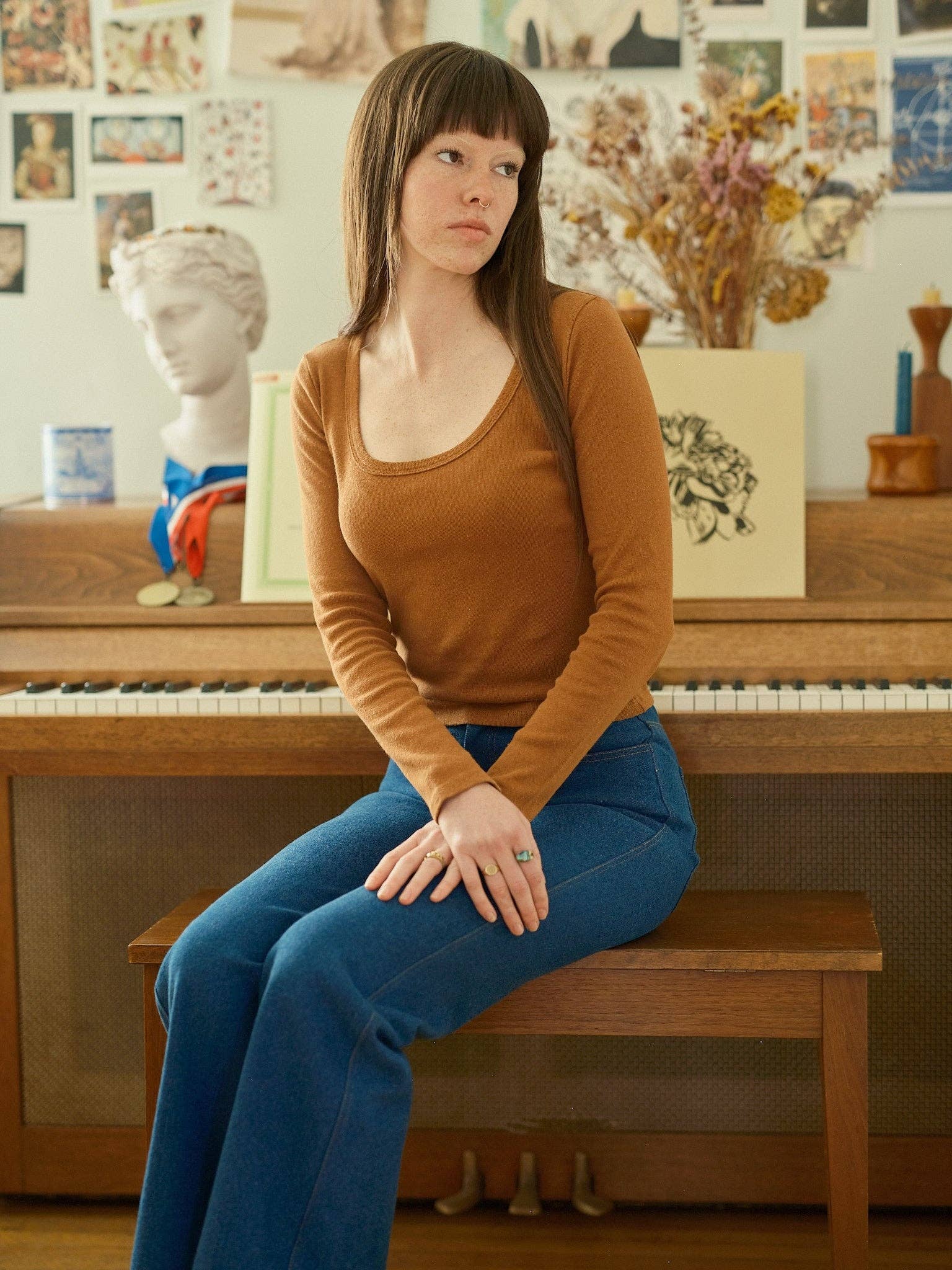 A woman with long brown hair sits on a piano bench, wearing the Mollusk Long Sleeve Rib Tee in brown and blue jeans. Behind her, art prints, a dried flower arrangement, and a white bust statue decorate the piano.