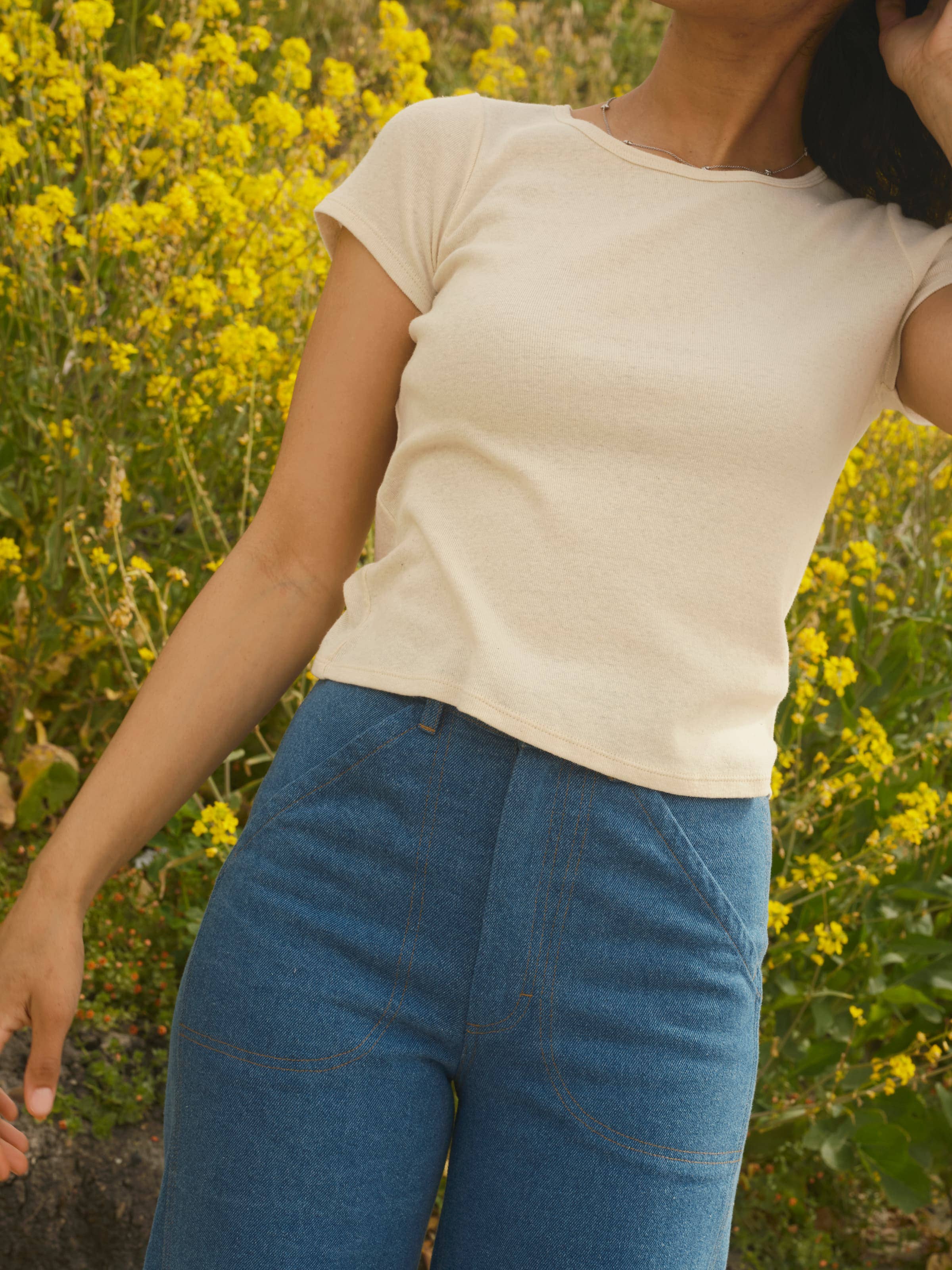 A woman stands outdoors before yellow wildflowers, wearing Mollusk Painter Pants in Indigo and a short-sleeve beige top, her head turned to the side and face partially out of frame.