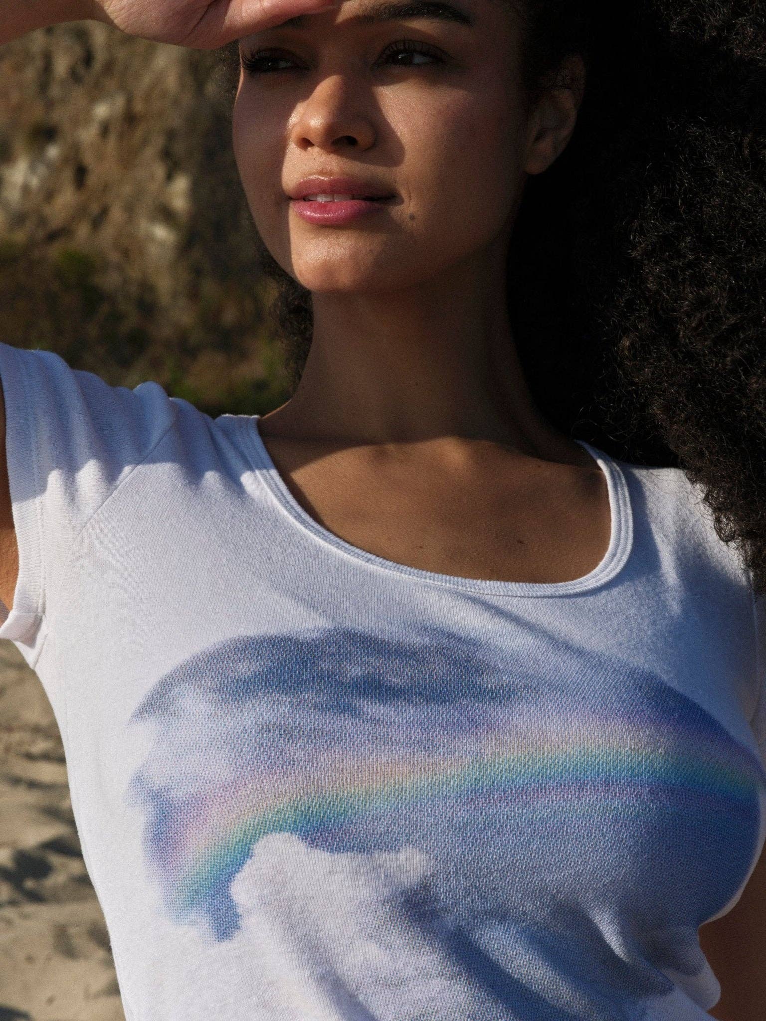 A woman outdoors partially shades her eyes as she wears the Mollusk CloudBreak Tee, made from sustainable hemp cotton with clouds and a rainbow across the chest. A blurred natural landscape forms the background.