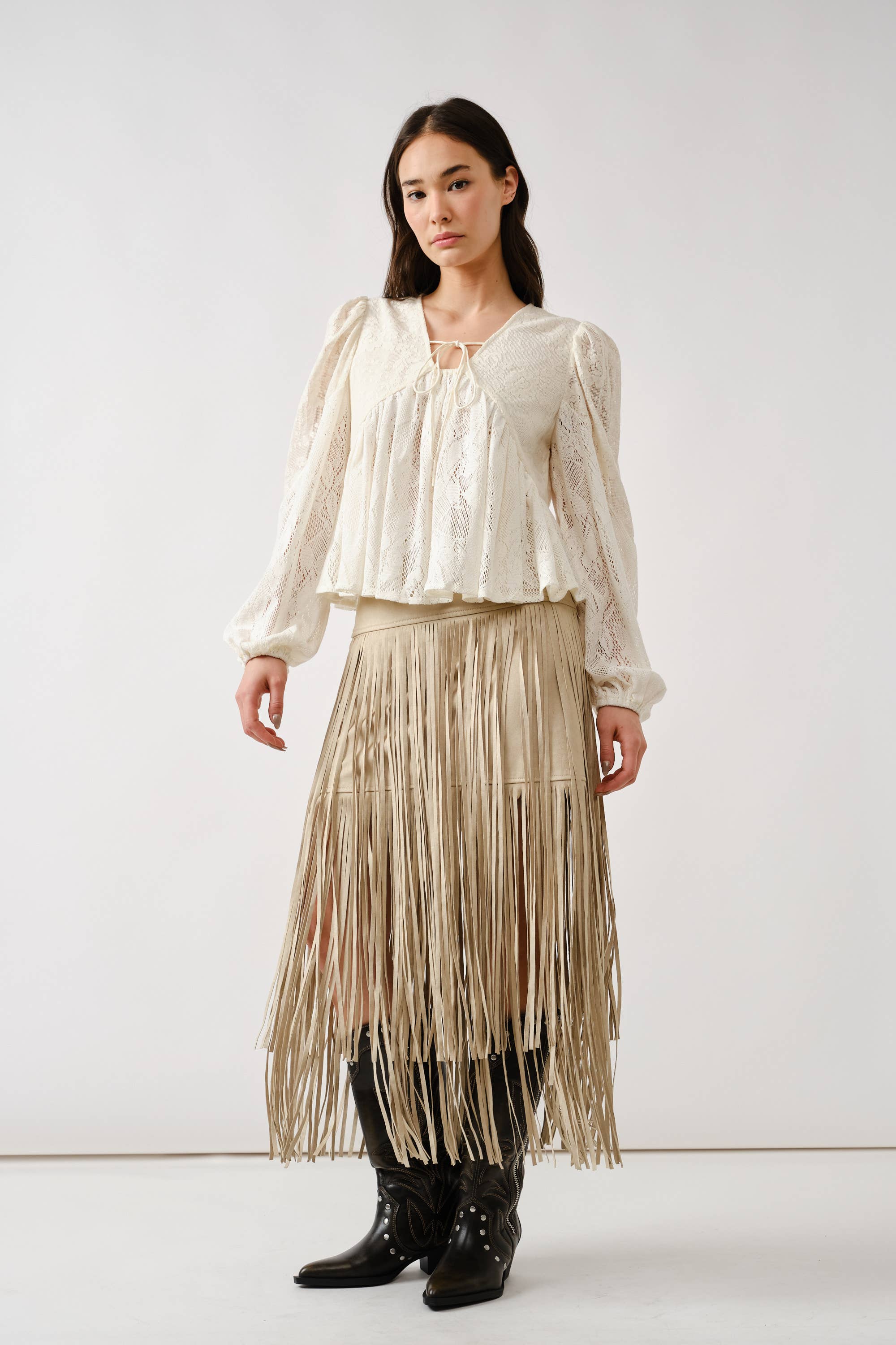 A woman with straight dark hair and a neutral expression wears the Aaron & Amber Long Sleeve Lace Babydoll top, paired with a beige long fringe skirt and black cowboy boots, standing against a plain background.