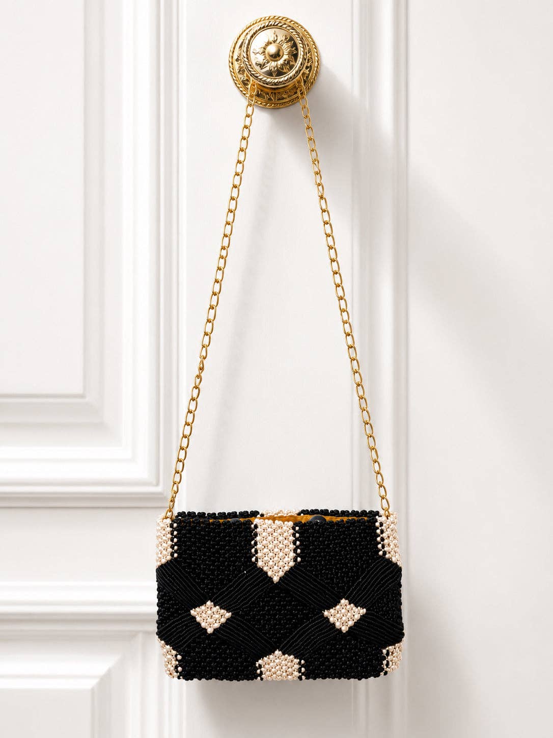 Coastal Knot Clutch hanging on ornate gold hook, featuring black and white beaded geometric design with gold chain strap