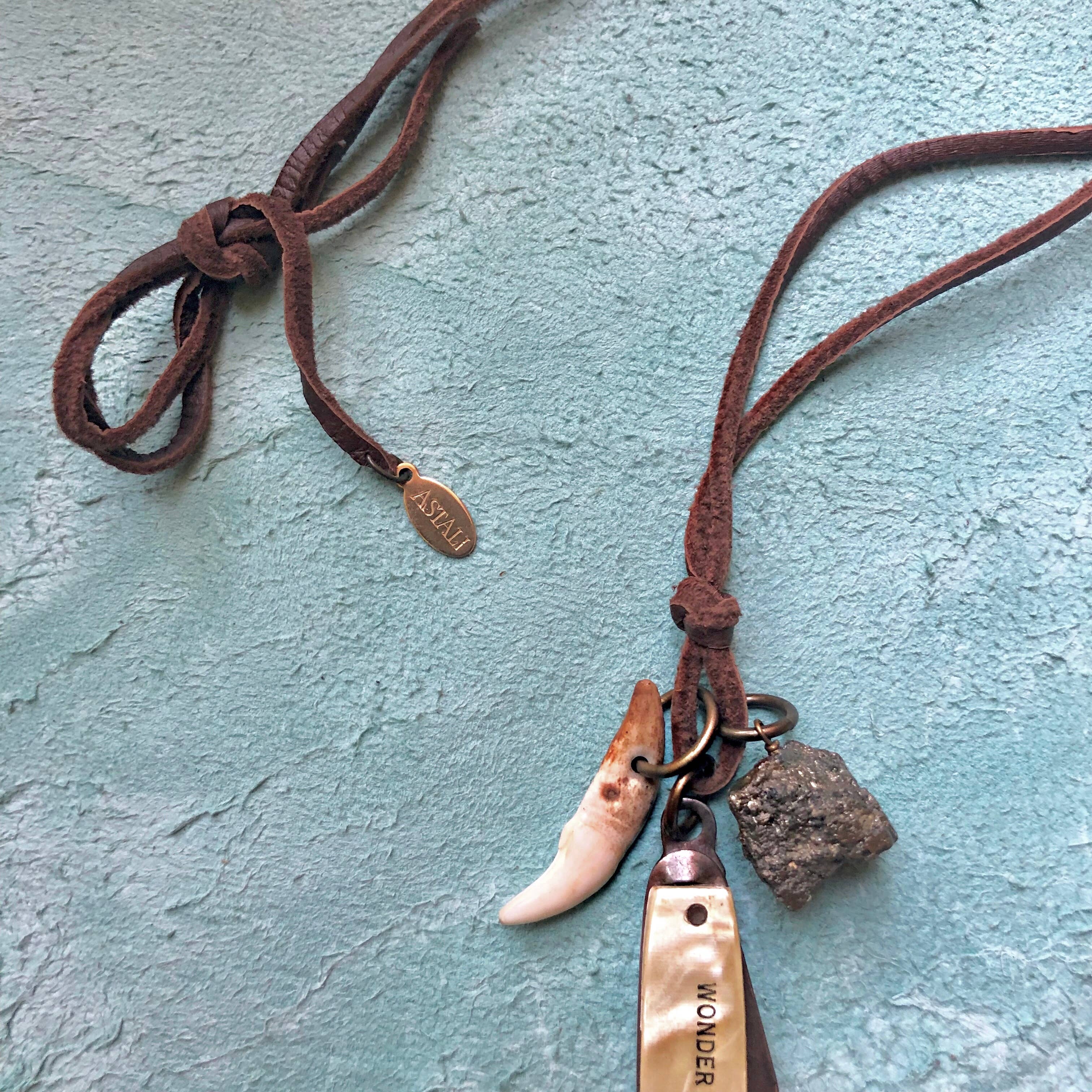 The ASTALI Vintage Pocket Knife & Coyote Tooth Necklace features pyrite and deerskin leather, accented with unique charms, resting on a textured blue surface.