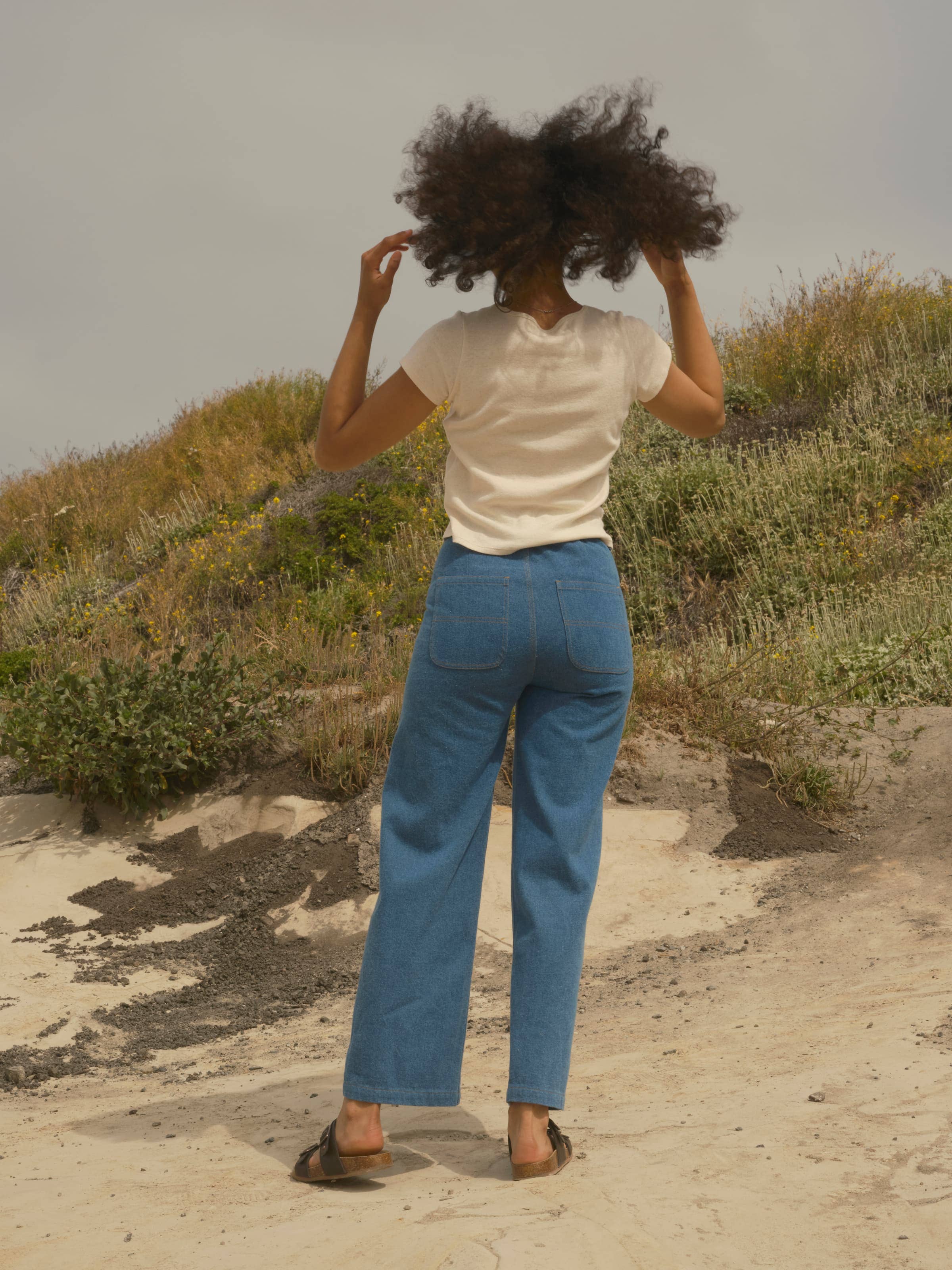 A person with curly hair, wearing a white tee, Mollusk Painter Pants in Indigo (100% cotton denim), and sandals stands outdoors on sandy ground with grassy hills behind them, facing away from the camera and adjusting their hair.