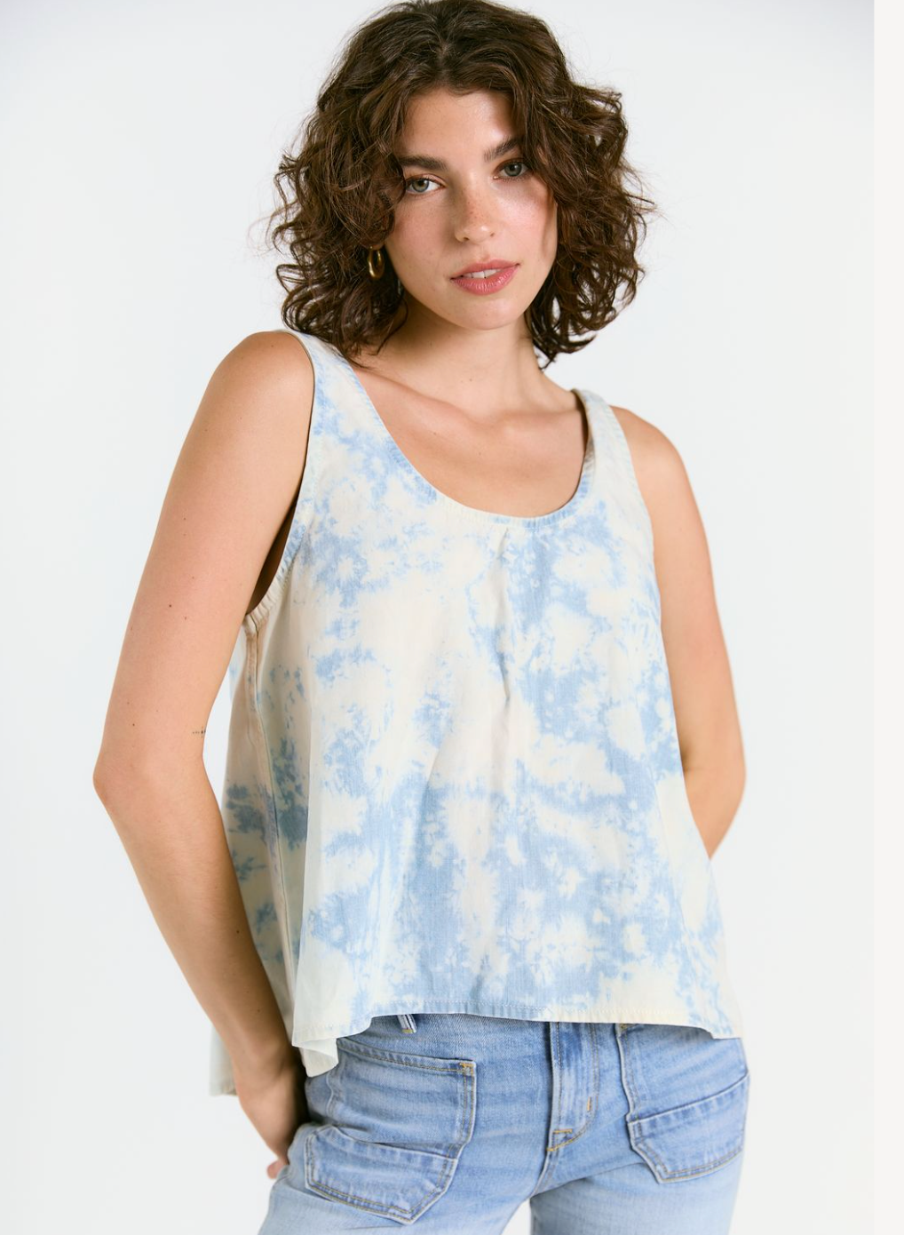 A woman with curly brown hair stands against a white background, wearing the WABI SABI TANK IN SKY TIE DYE by Porter Blue Apparel and light blue jeans, looking at the camera with a neutral expression.