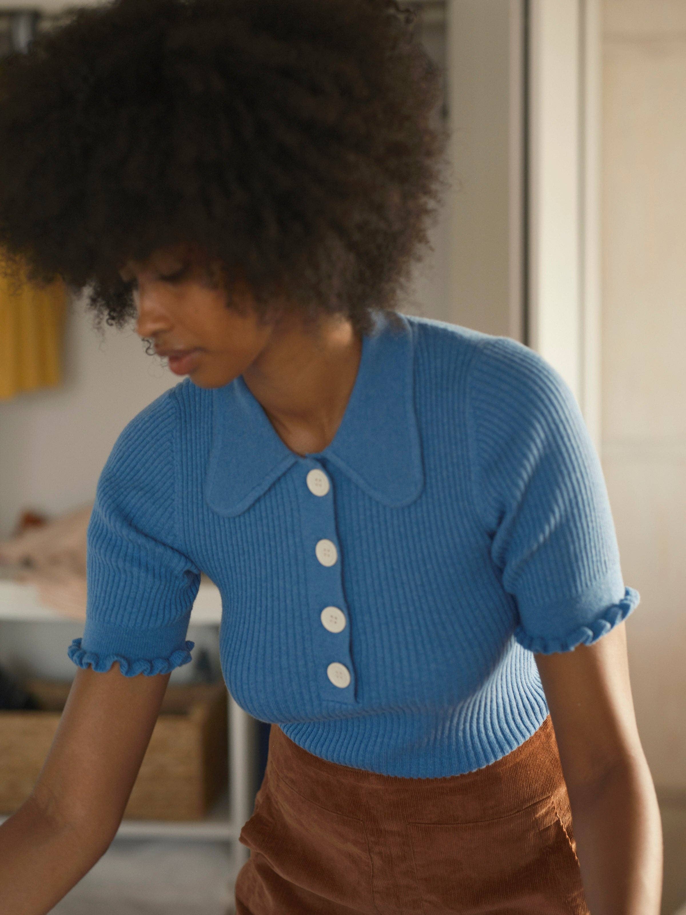 A person with curly hair wears the Mollusk Belle Tee—a blue, short-sleeved, vintage-inspired ribbed knit top with a large Peter Pan collar and white buttons—paired with brown pants while standing indoors and looking down.