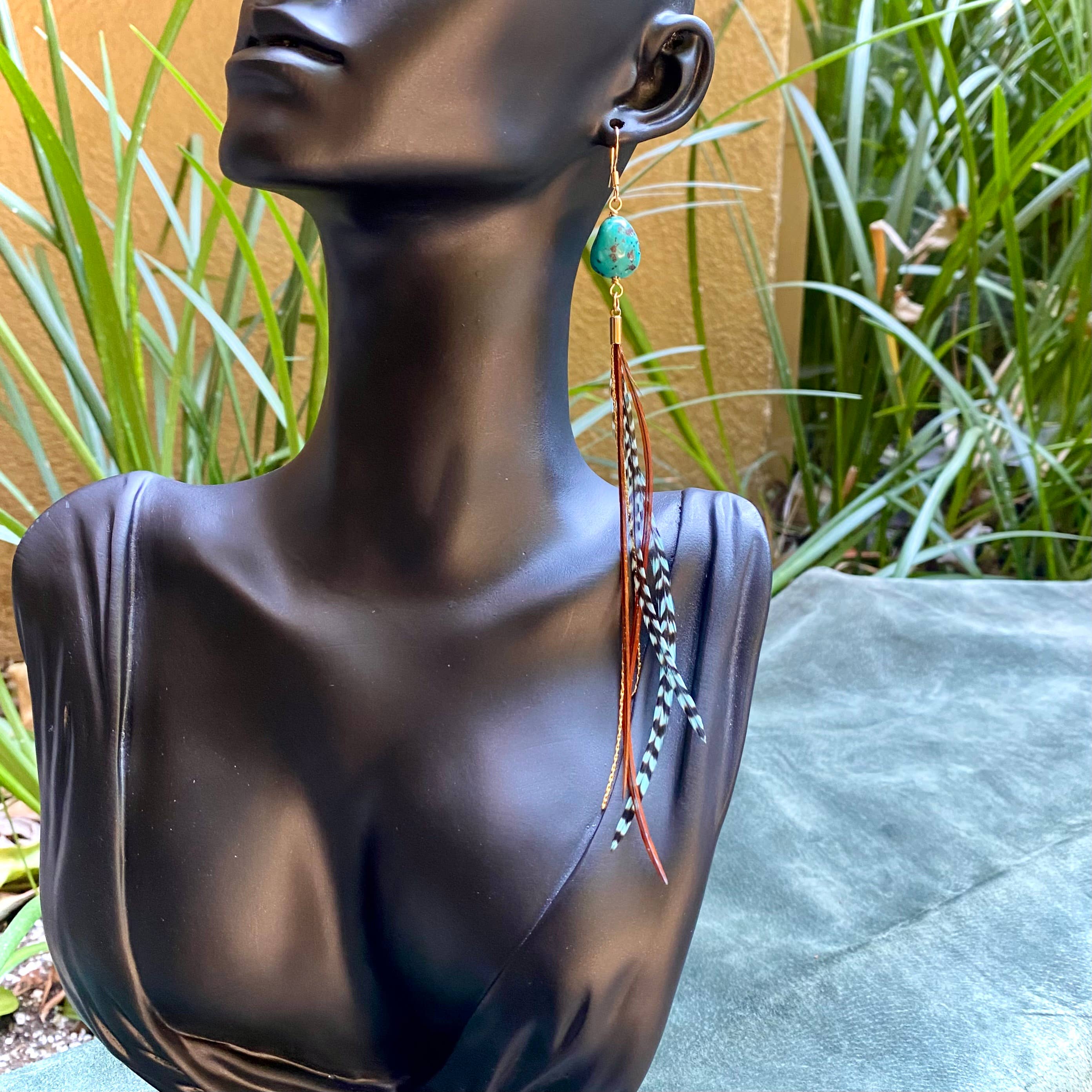 A black mannequin bust showcases ASTALI’s Turquoise & Mini Feather Earrings—Teal Grizzly & Fawn/Gold—handcrafted with turquoise stones, teal grizzly and fawn feathers, and gold-plated accents. Green plants and a tan wall emphasize their unique style.