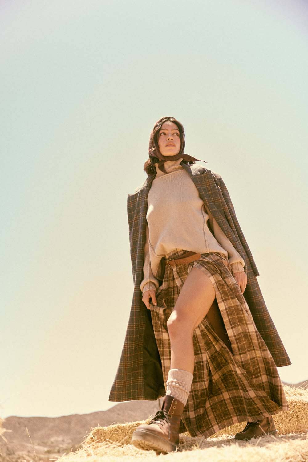 A woman stands outdoors on dried grass, wearing the Promesa USA Plaid Felt Double-Breasted Calf-Length Overcoat over a cream sweater and plaid high-slit skirt, with socks and boots. The clear sky and distant mountains form the backdrop.
