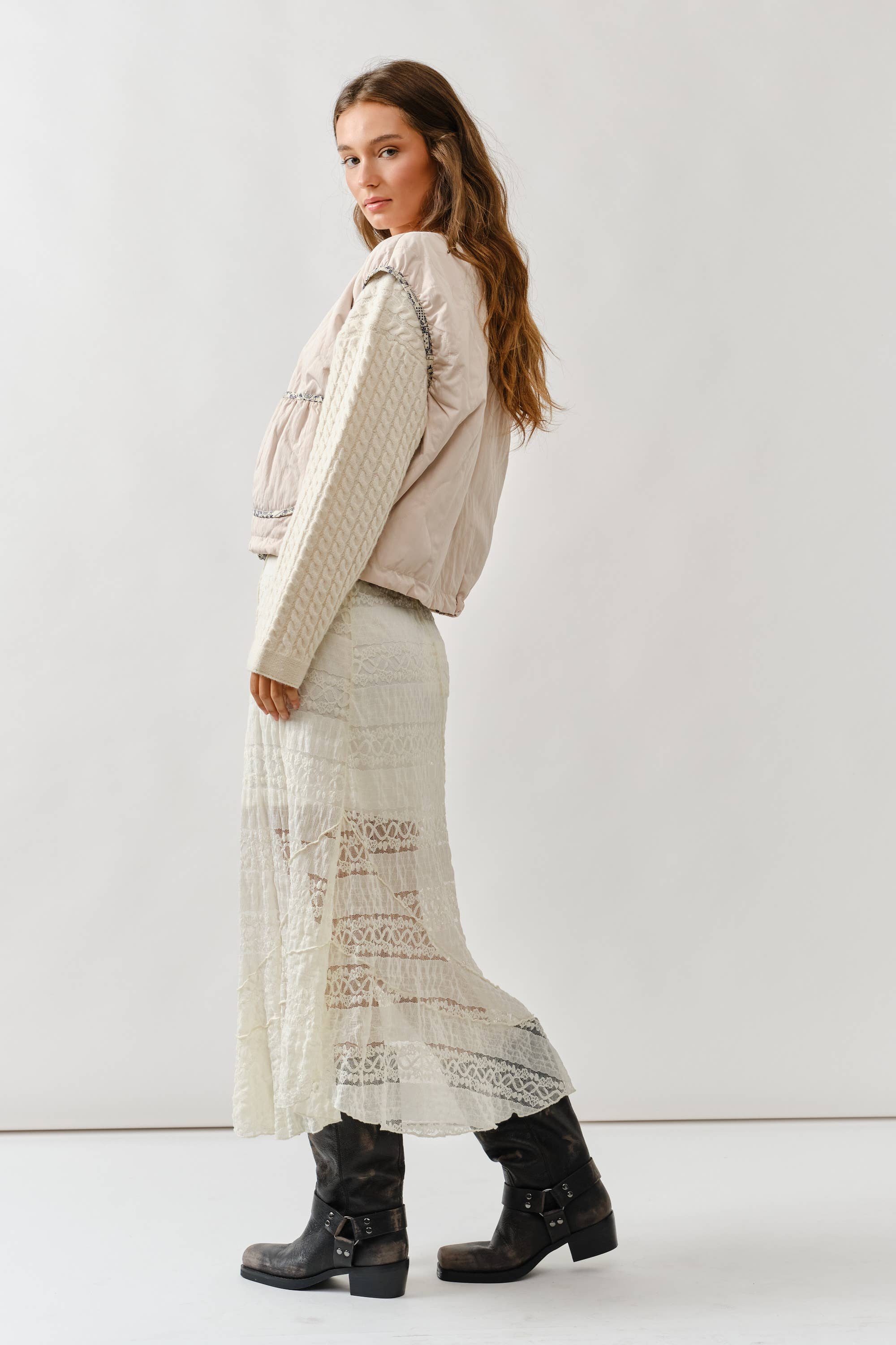A woman with long wavy hair models the Aaron & Amber Contrast Detail Lightweight Vest over a cream lace midi skirt and black boots, posing sideways against a plain white background.
