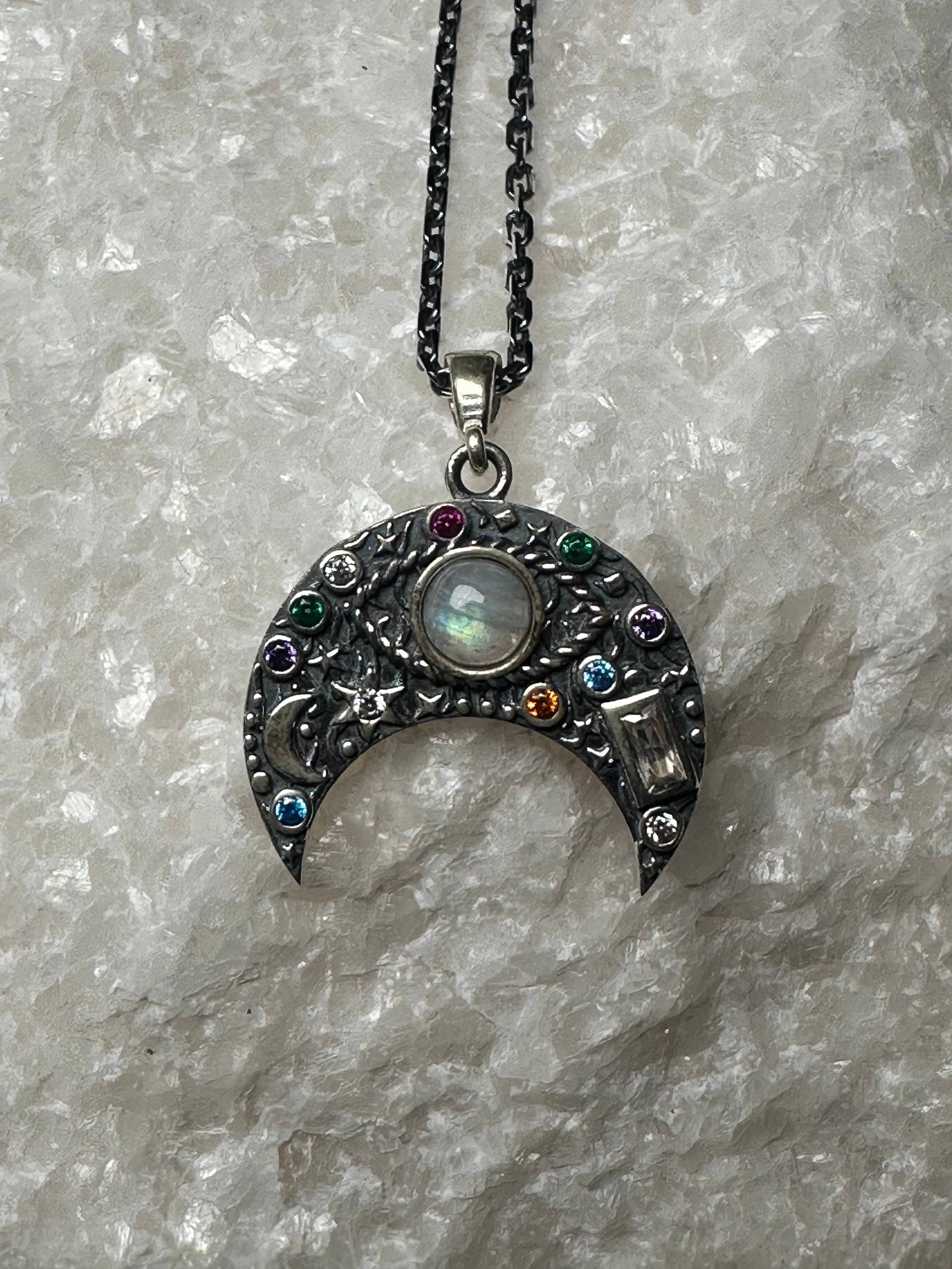 The Flesh & Soul CRESCENT MOON MOONSTONE Evil Eye Sterling Silver Necklace features a central round stone and colorful gems on a black chain, shown against textured white crystal.