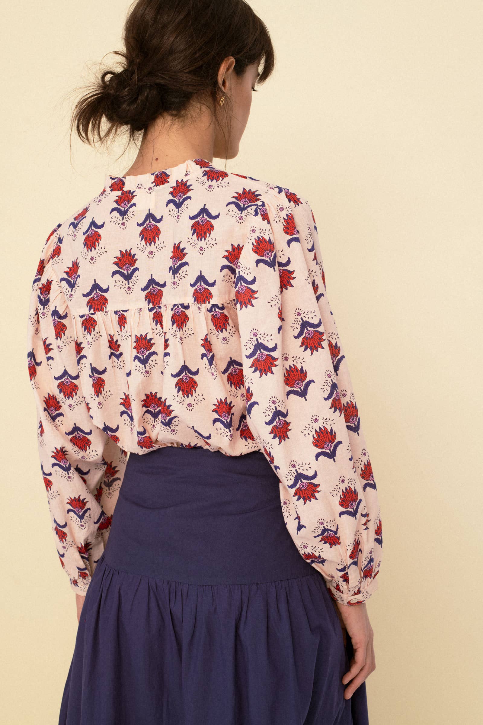 A woman with dark hair tied back is facing away, wearing the Sonmer Shop USA Vanessa Blouse Peach— a long-sleeved cotton voile blouse with red, purple, and blue florals on a light background—paired with a high-waisted purple skirt.