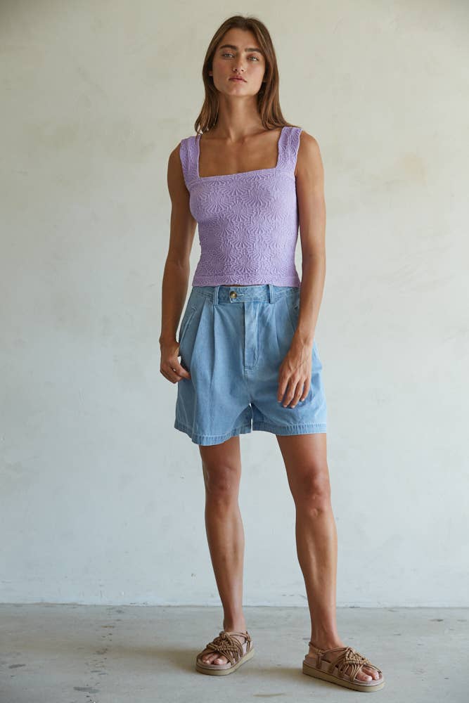 A woman with straight brown hair and a neutral expression stands against a plain light wall, wearing the By Together Knit Modal Nylon Spandex Seamless Jacquard Tank in lavender, light blue high-waisted shorts, and tan sandals.