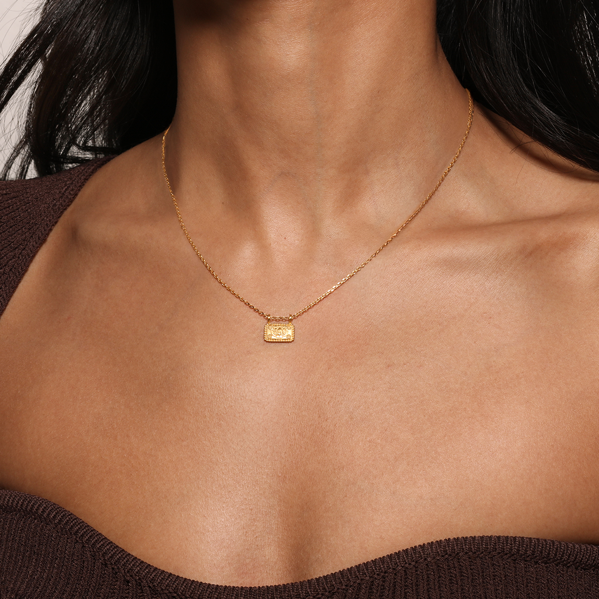 A woman wears the Satya Jewelry 16” Lotus Plate Necklace, featuring a gold-plated rectangular lotus pendant. She's dressed in a brown off-shoulder top, drawing attention to her neck and collarbone—a symbol of new beginnings.