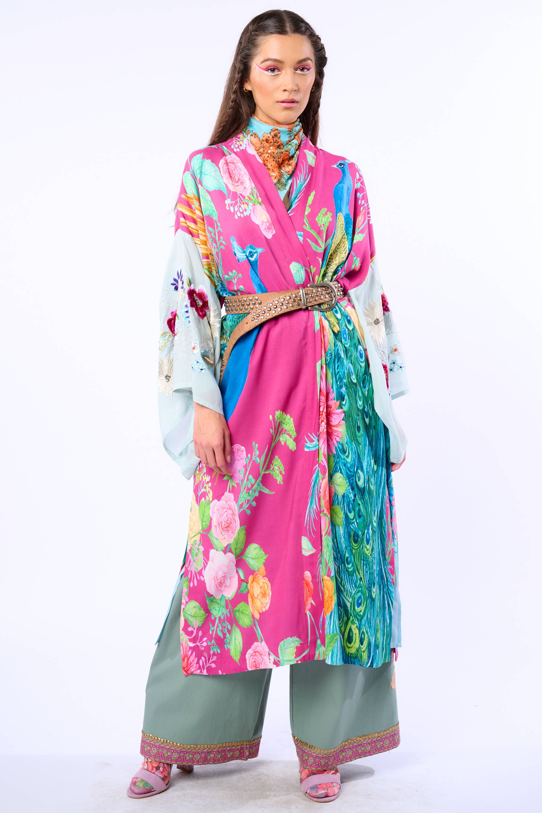 A woman poses against a white background in the Marie Antoinette Kimono by Aratta, featuring embroidered floral sleeves and a belted waist, styled over wide green pants with embroidered cuffs. She wears pink eyeshadow and has long, wavy hair.