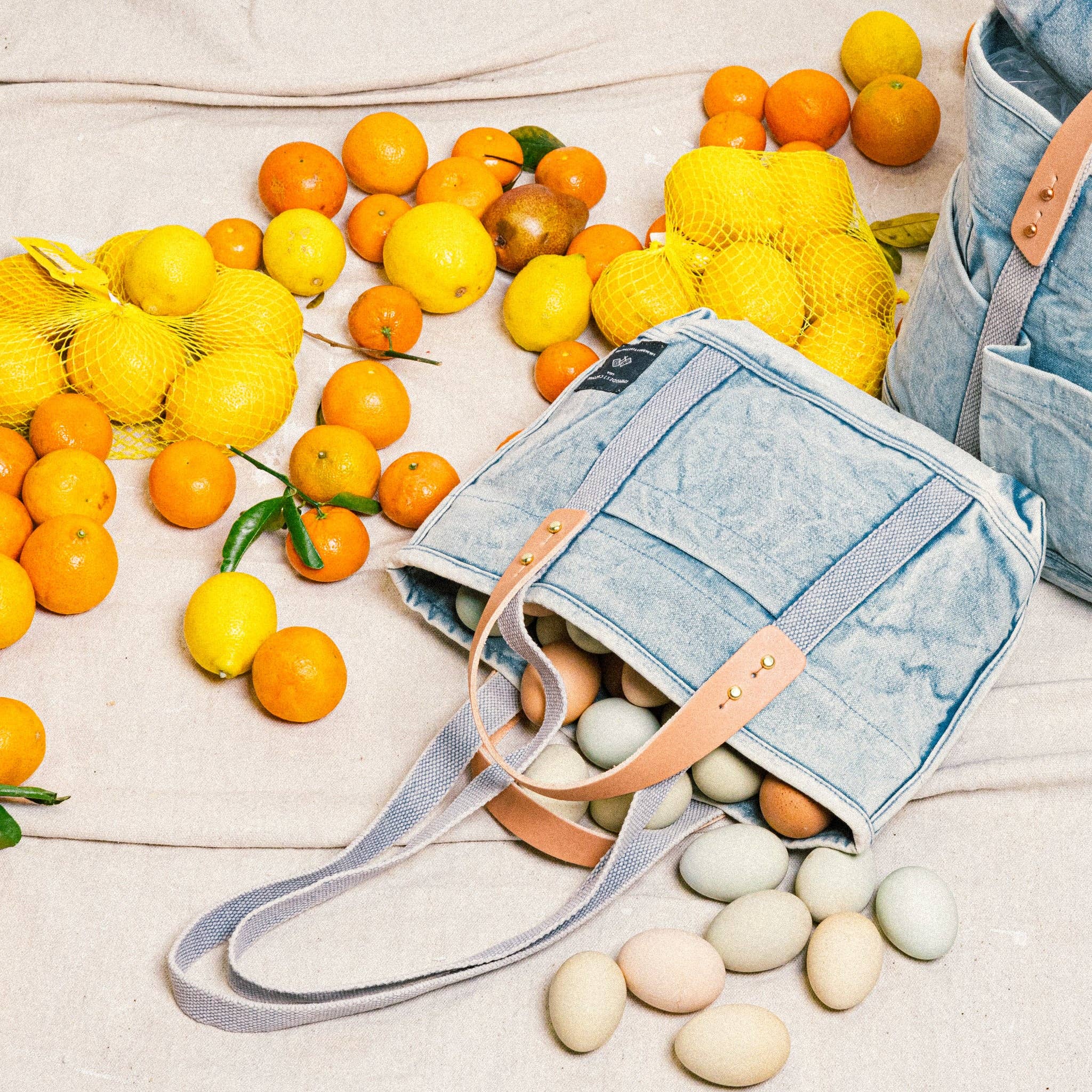 Lunch Tote | Acid Wash canvas bag with leather handles surrounded by citrus fruits and pastel eggs on fabric surface