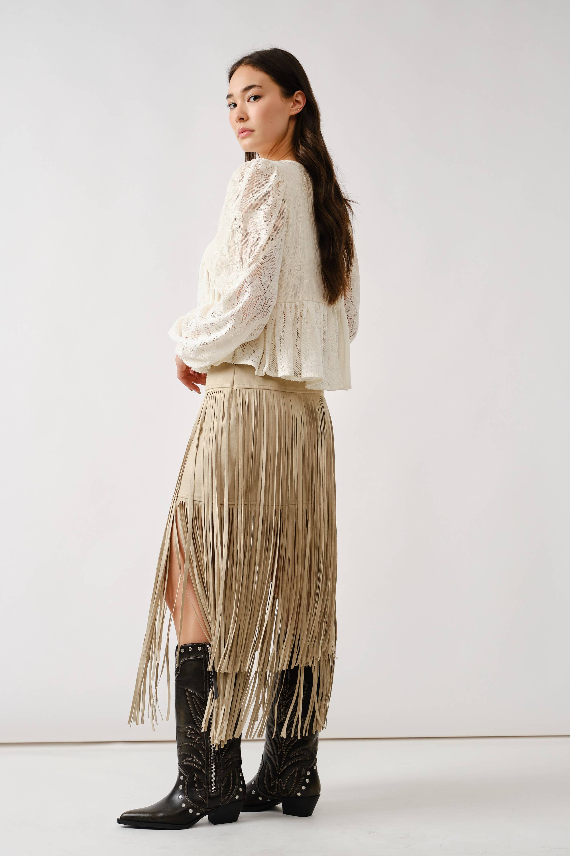 A woman stands sideways against a white background, wearing the Aaron & Amber Long Sleeve Lace Babydoll—a romantic white floral lace blouse—paired with a beige fringed skirt and black cowboy boots.