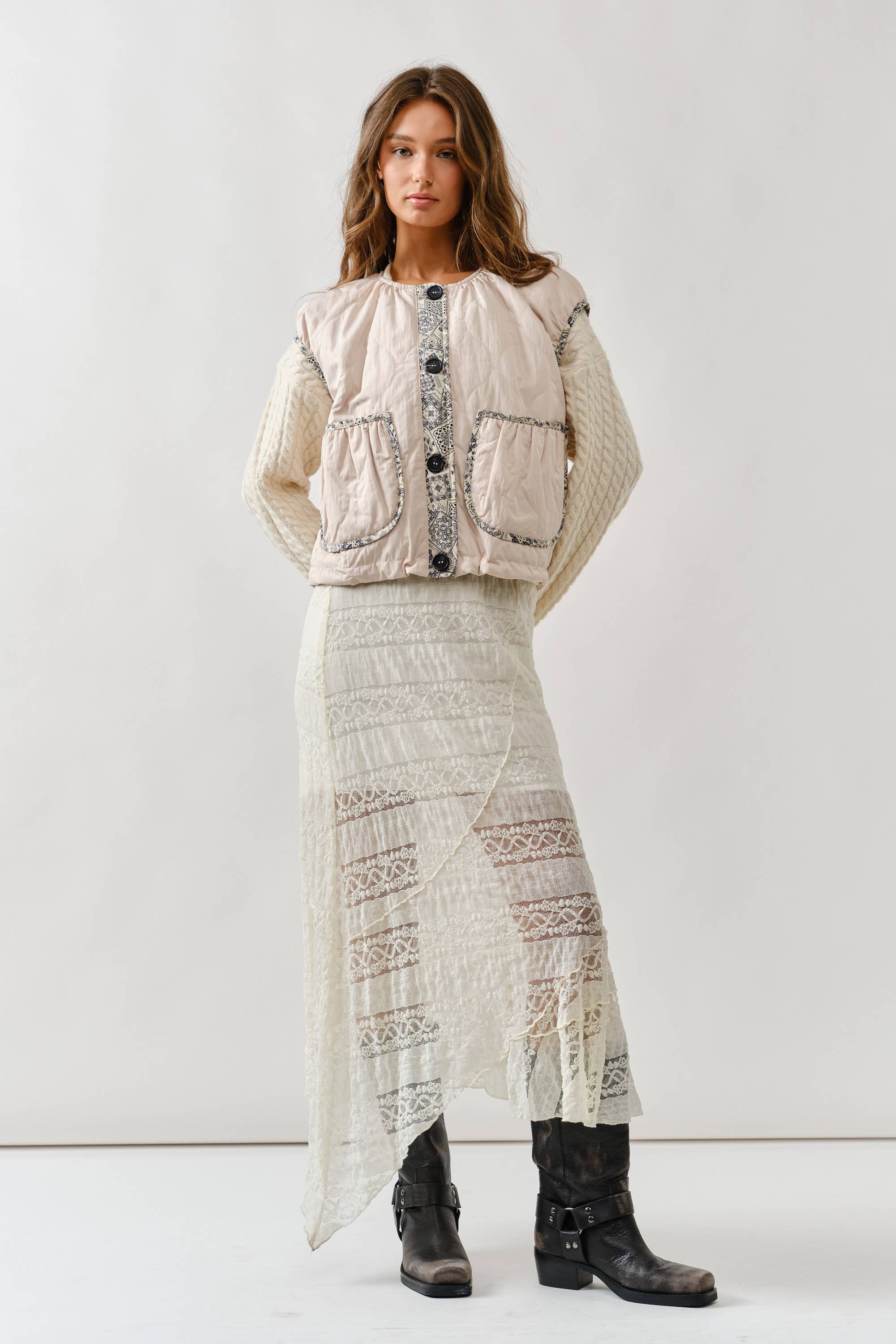 A woman with long wavy hair and a neutral expression stands against a plain background, wearing a textured cream sweater, the Aaron & Amber Contrast Detail Lightweight Vest, a long asymmetrical lace skirt, and black boots.