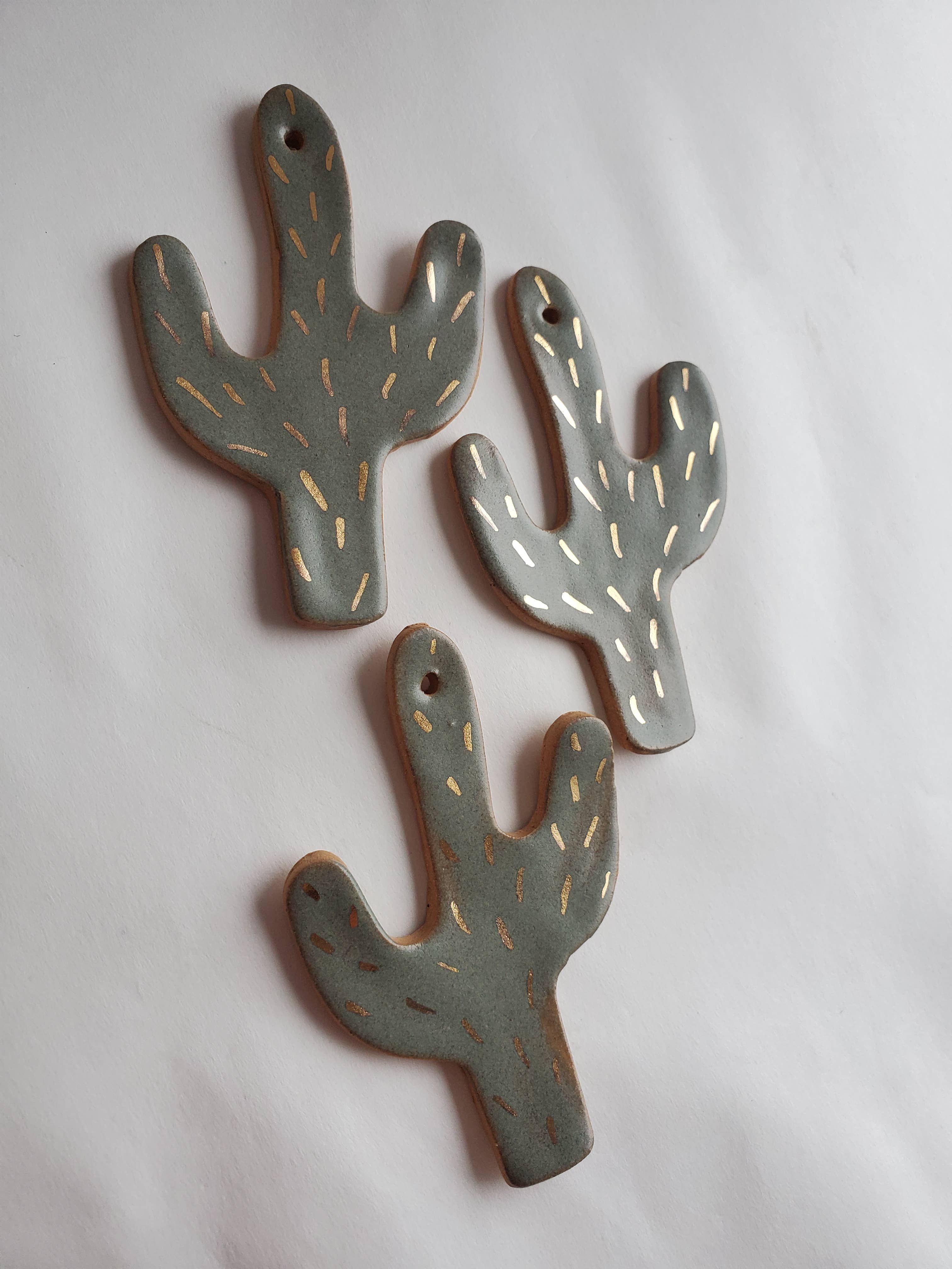 Three handmade ceramic Cactus ornament pieces in muted green with subtle gold highlights on a white surface