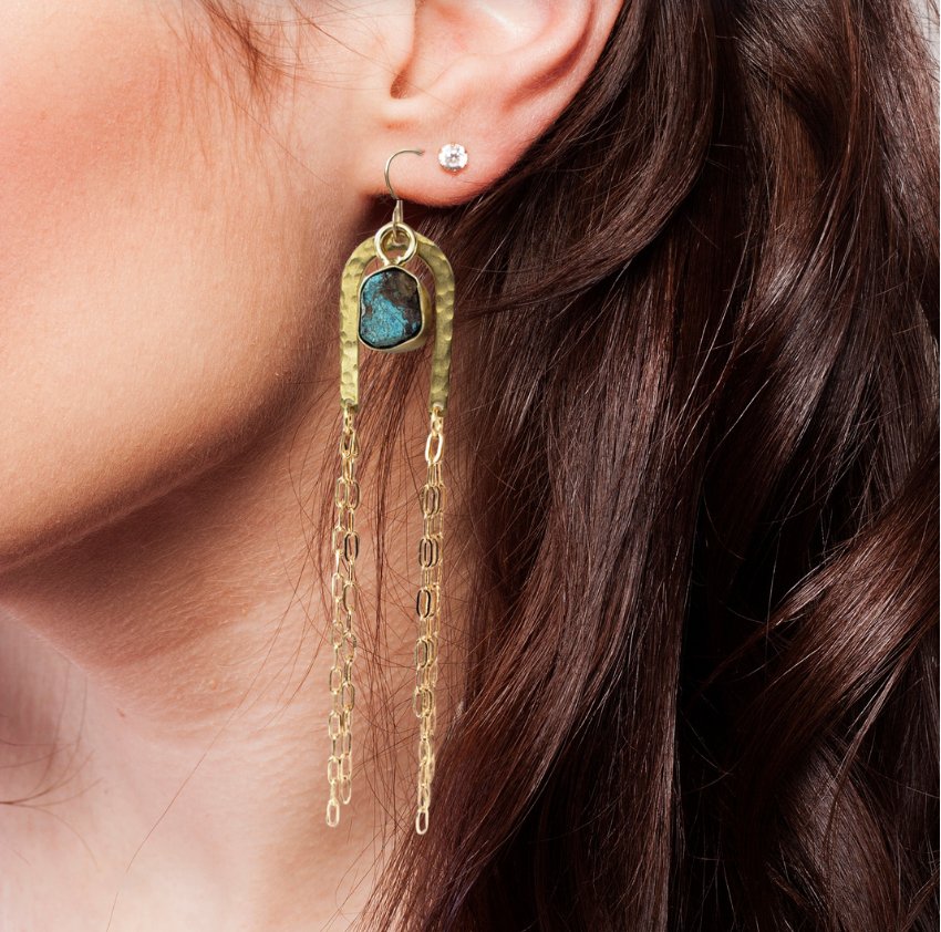 Close-up of a womans ear wearing Dreamers of Dreams Gold Rainbow Arch Chain Duster Earrings, with blue chrysocolla pendants and 14K gold fill chains, shown against her dark wavy hair.