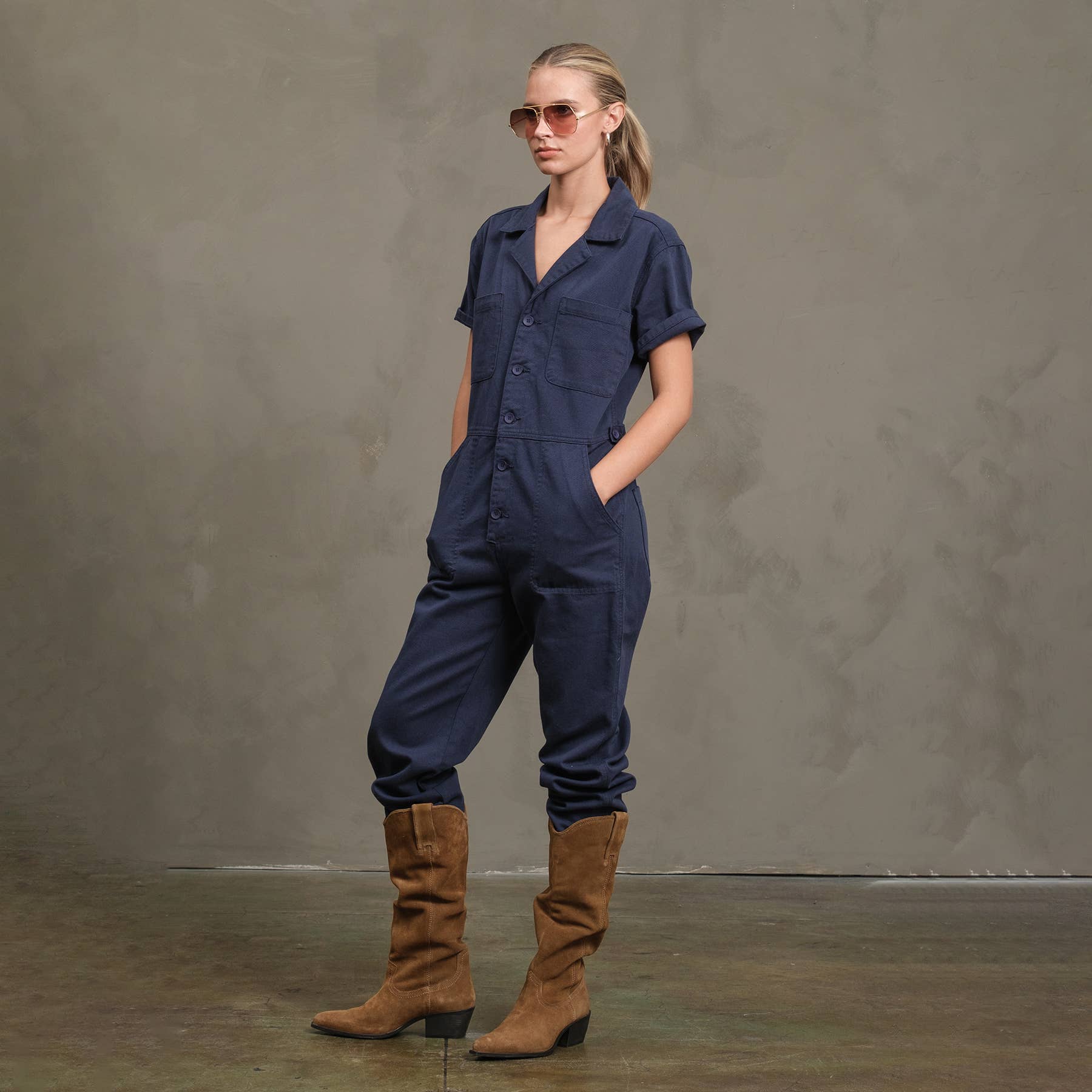 A woman in the RAISONNEL Field Jumpsuit (RJP1001), brown suede boots, and sunglasses stands with her hands in her pockets against a gray textured background.