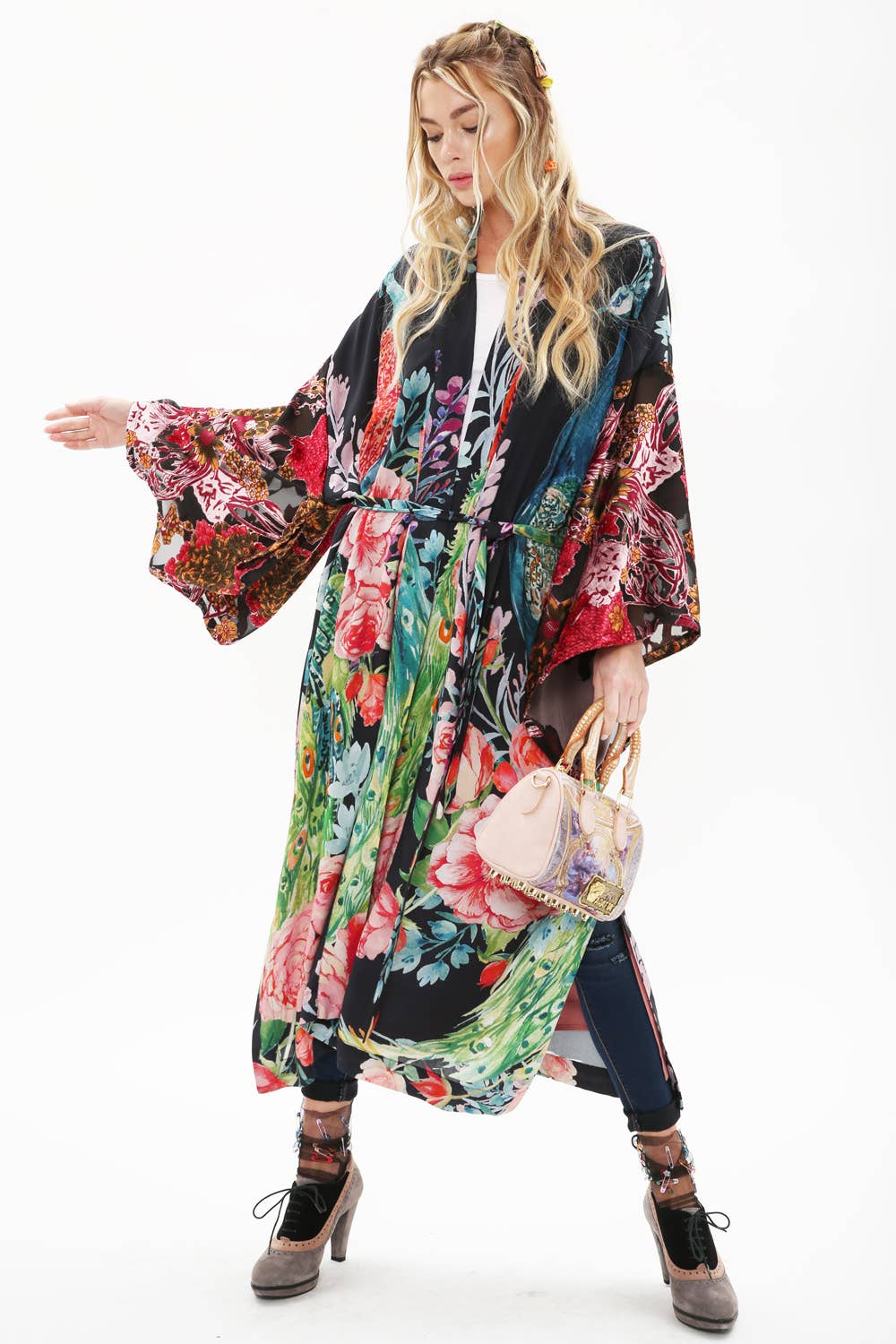 A woman poses against a white background wearing the Aratta Sweet Fantasy Kimono with burnout velvet sleeves, paired with dark pants, grey heels, a pink handbag, and her long wavy hair styled in loose braids.