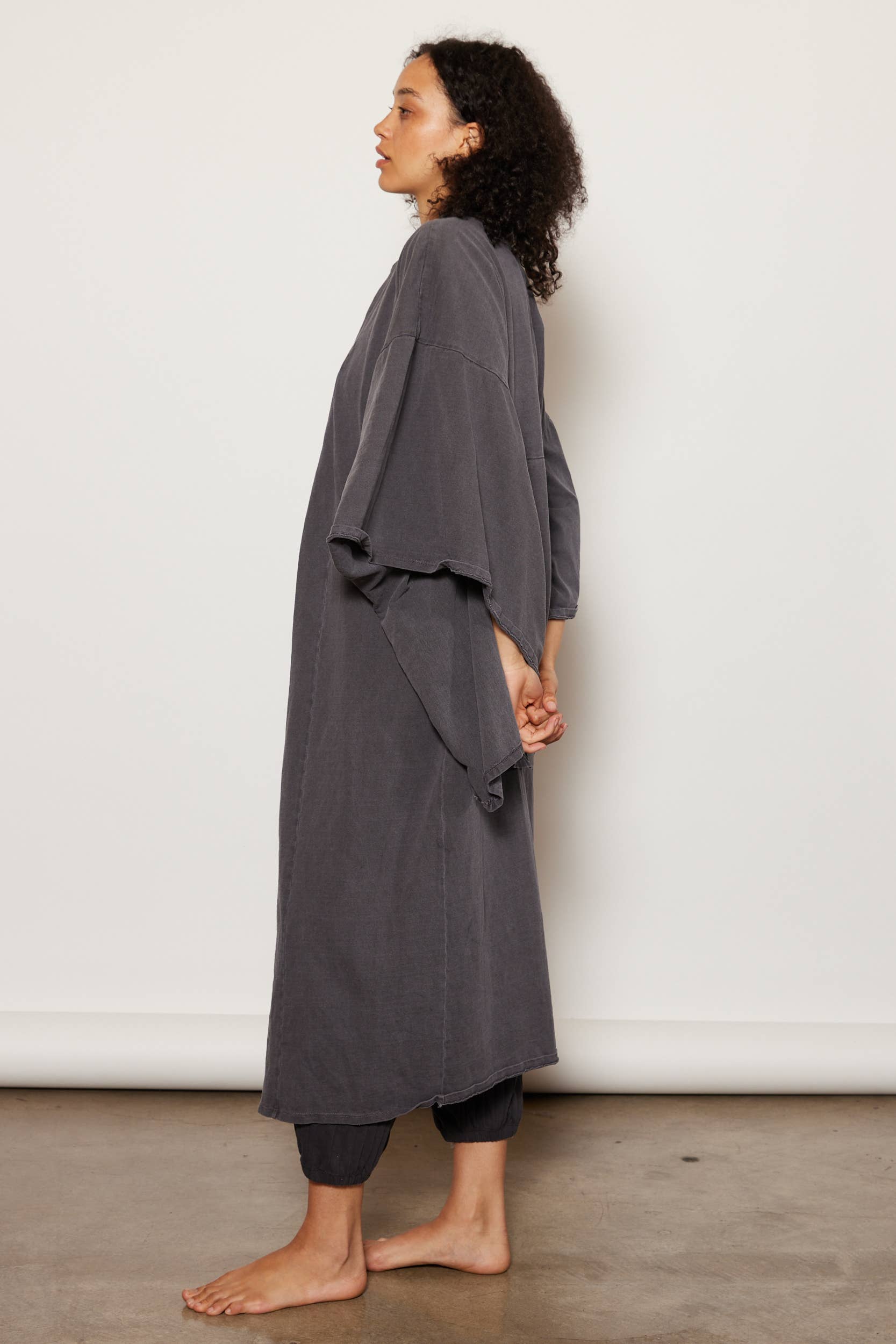A person with curly hair stands barefoot in profile against a plain background, wearing People Of Leisure’s oversized gray organic cotton Dawn Robe with wide sleeves over matching pants. Ethically made in Los Angeles.