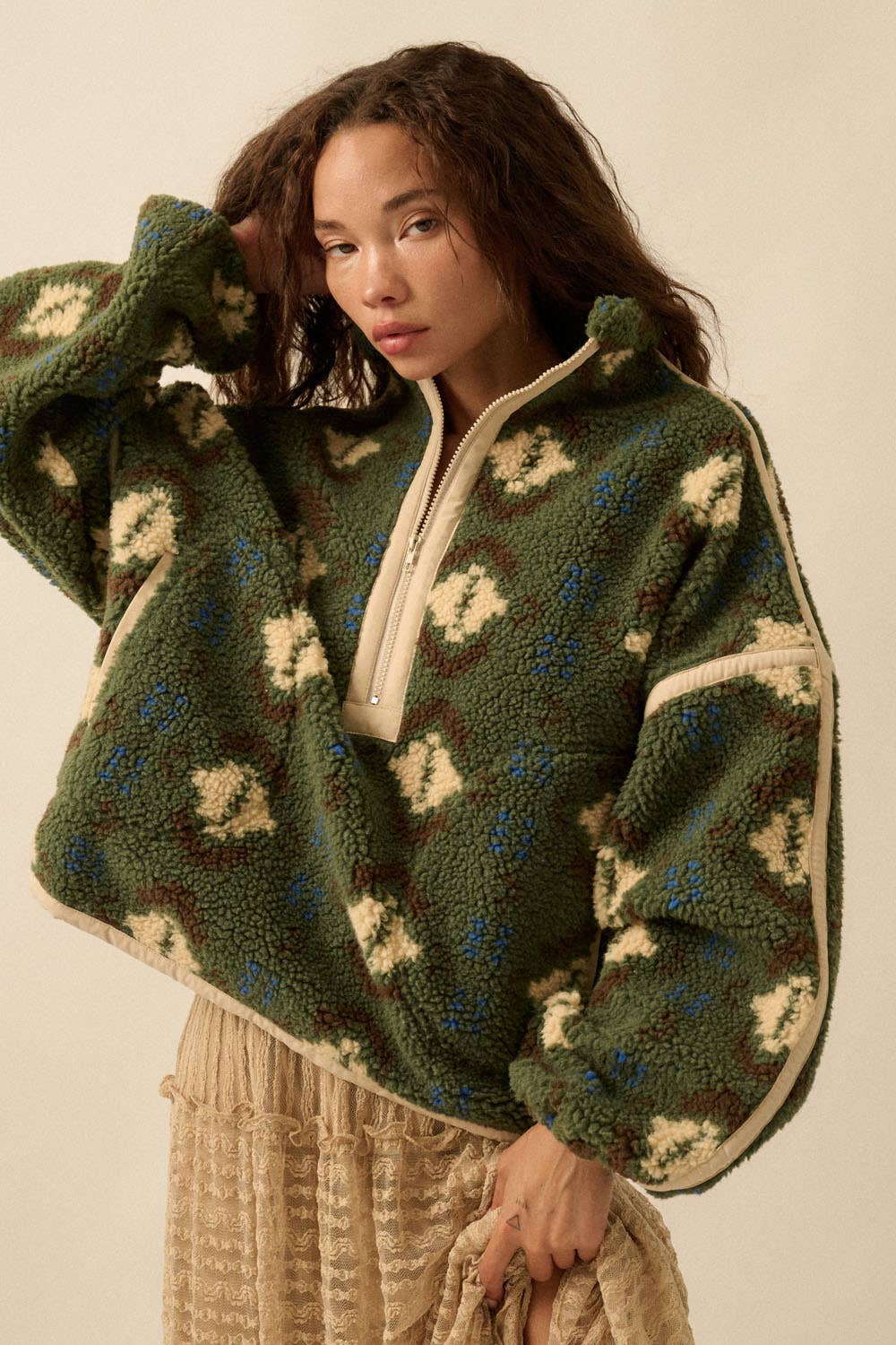 A woman poses against a neutral background, wearing the Promesa USA Geo-Print Sherpa Fleece Half-Zip Pullover Jacket in green and cream with beige trim, paired with a light beige textured skirt, and touches her hair.