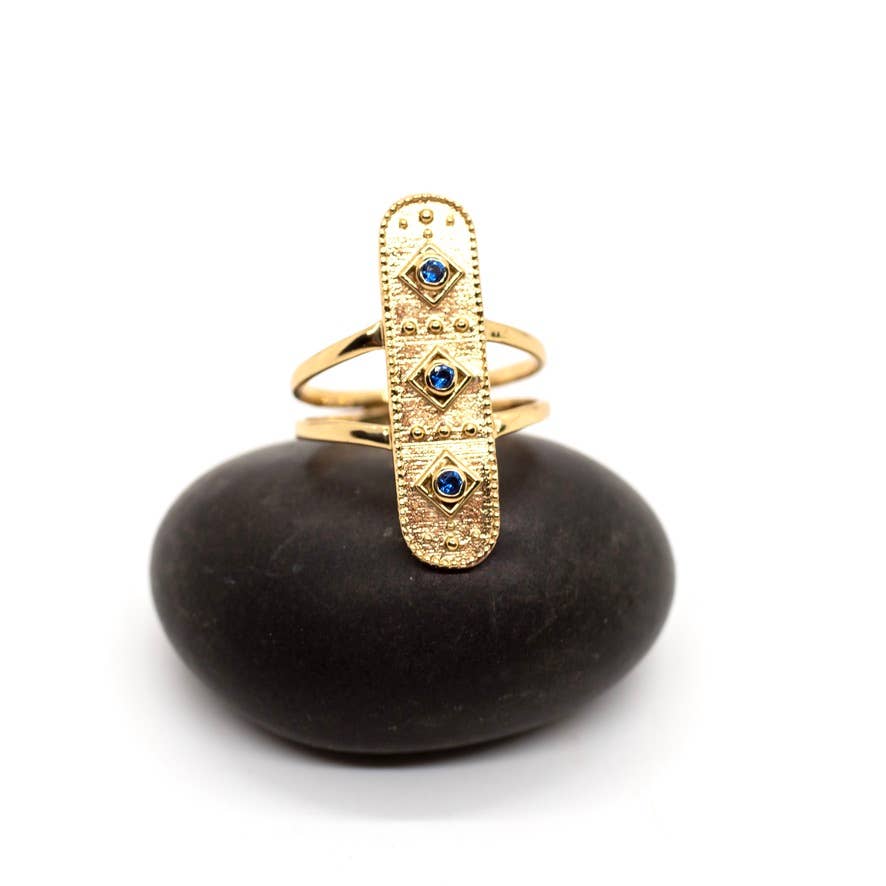 The Baizaar Sapphire Healer Ring features a textured oval face with three faceted sapphire stones, shown upright on a smooth black stone against a white backdrop.