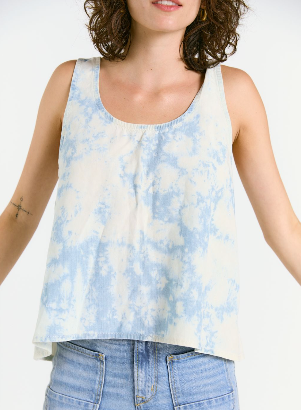 A person in the Porter Blue Apparel WABI SABI TANK IN SKY TIE DYE and light blue denim jeans stands with arms slightly open; their face is partially out of frame.