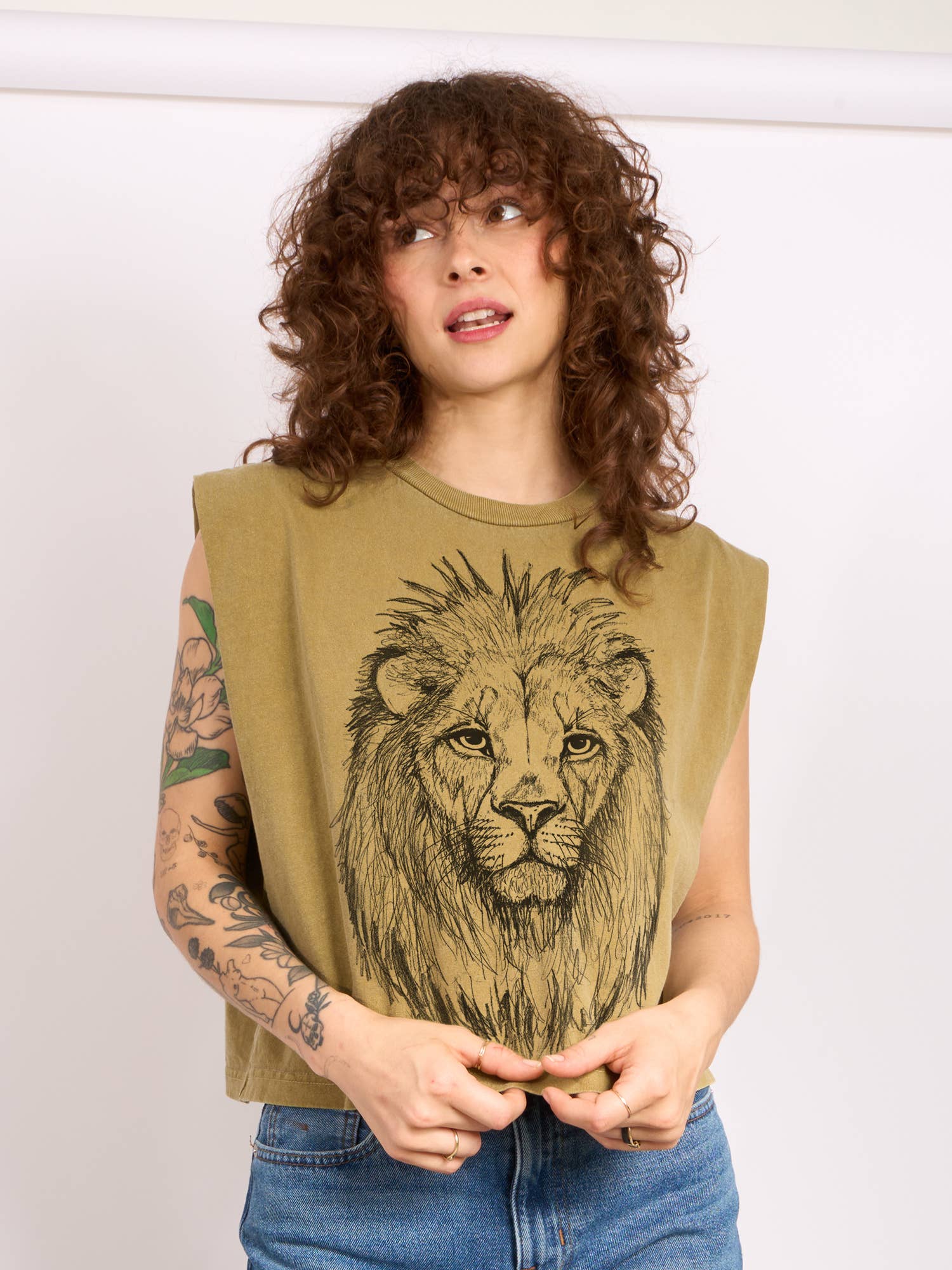 A person with curly hair and tattoos wears the Supermaggie @ Faire Lion Maisie Heavyweight Muscle Tee, a boxy cotton top featuring a detailed lion’s face, paired with blue jeans against a plain light background.