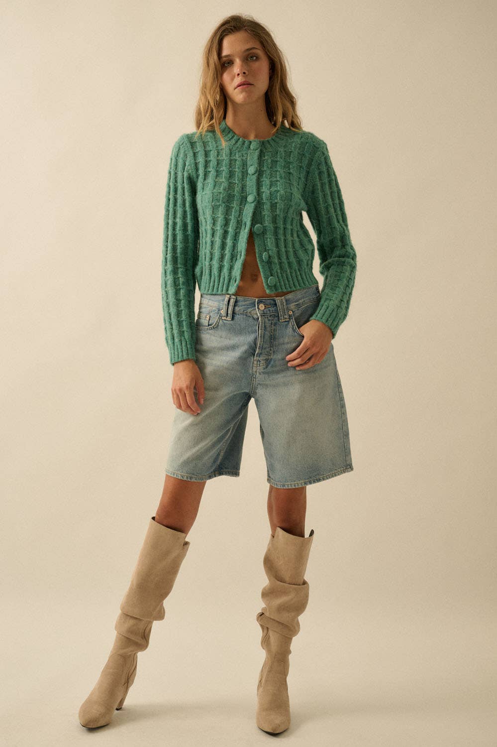 A woman poses confidently in a Promesa USA Cable Knit Button-Up Cardigan Sweater, paired with light blue knee-length denim shorts and beige slouchy knee-high boots. She stands against a light background with loose hair and one hand in her pocket.