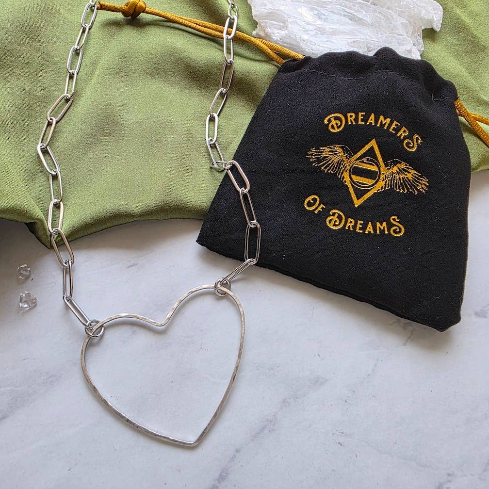 The Big Silver Heart - Paperclip Chain Necklace by Dreamers of Dreams, featuring a hammered heart charm, rests on a marble surface beside a black Dreamers of Dreams pouch, with a green cloth and crystals visible.