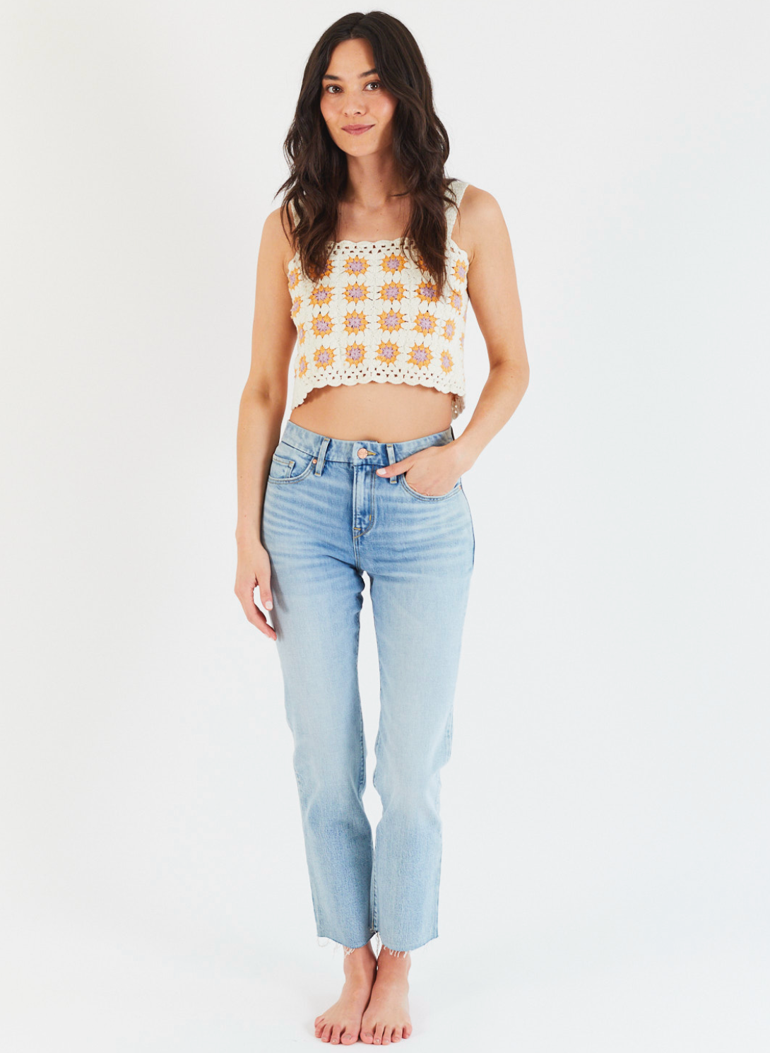 A woman with long dark hair wears Porter Blue Apparels REBEL STRAIGHT IN EDEN WASH jeans and a crocheted crop top. She stands barefoot against a white background, smiling softly with one hand in her pocket.