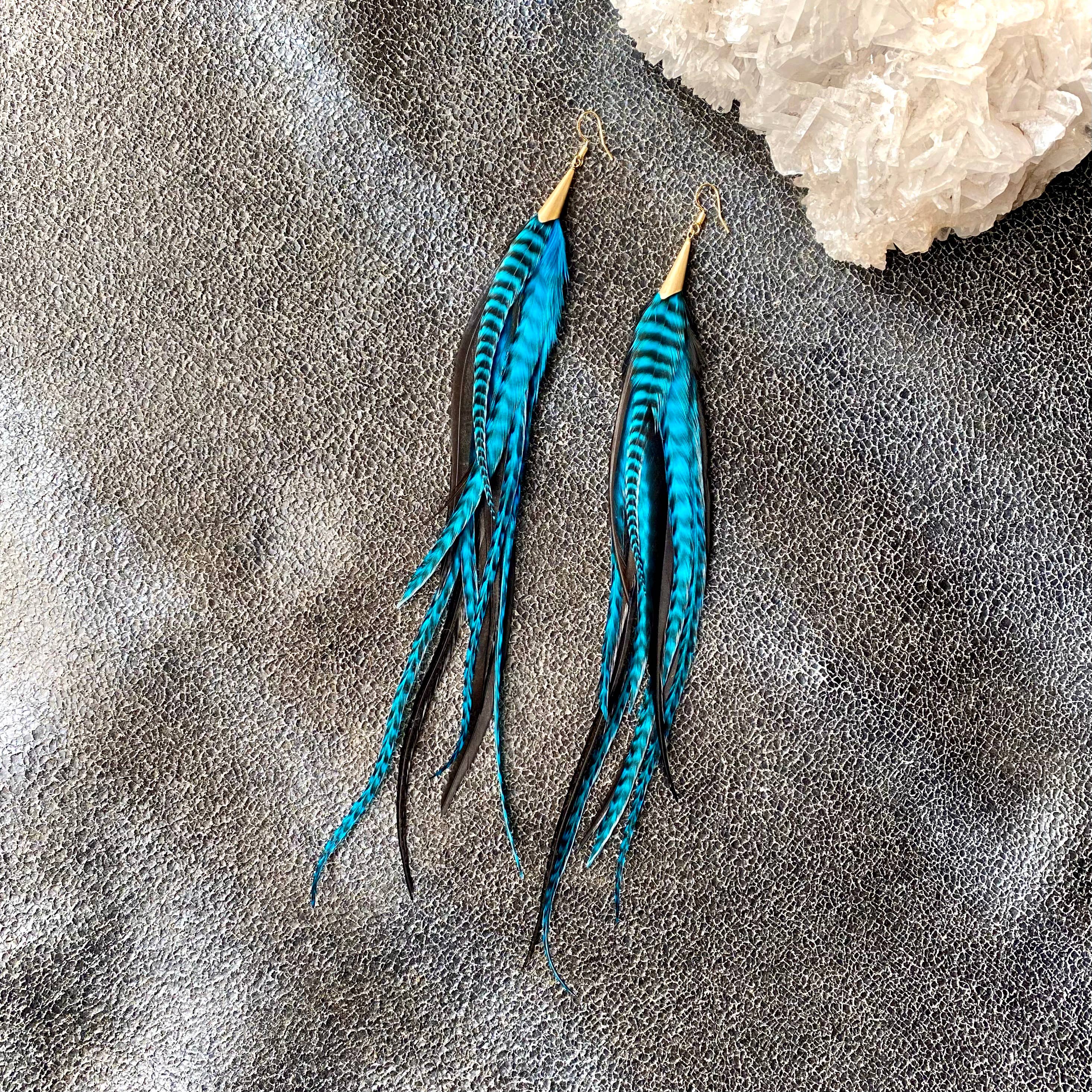 ASTALIs Long Feather & Cone Earrings in Turquoise Grizzly and Black/Gold feature vibrant feathers and gold-tone hooks, displayed on a textured gray surface beside a white crystal cluster.