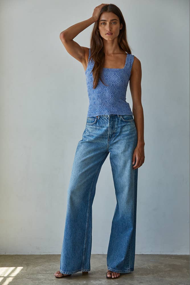 A woman stands indoors against a plain wall, wearing the By Together Knit Modal Nylon Spandex Seamless Jacquard Tank and high-waisted, wide-leg blue jeans; one hand touches her head while the other rests by her side.