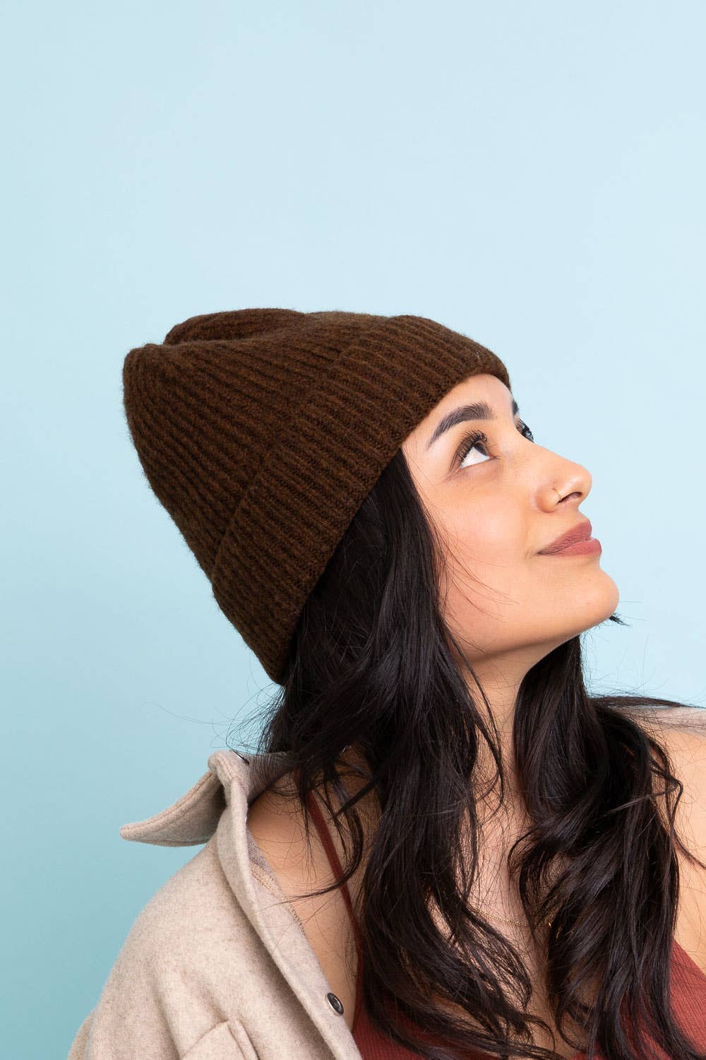 A woman with long dark hair wears the Versatile Cuffed Beanie – Cozy Rib Knit Hat by Leto Accessories in brown, paired with a beige jacket and reddish top, smiling slightly against a solid light blue background.