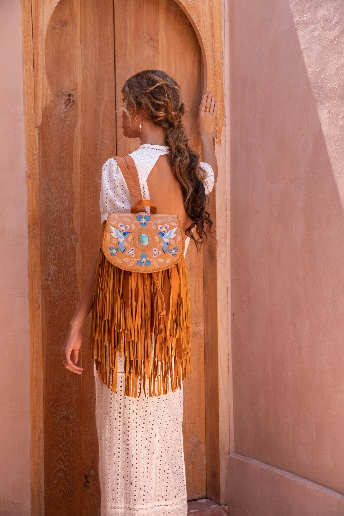 A woman in a white eyelet dress faces a wooden door, wearing the Jodi Lee Monterey Backpack in Antique Tan—crafted from cow leather with blue and white floral embroidery. Her wavy hair is tied back as she touches the door.