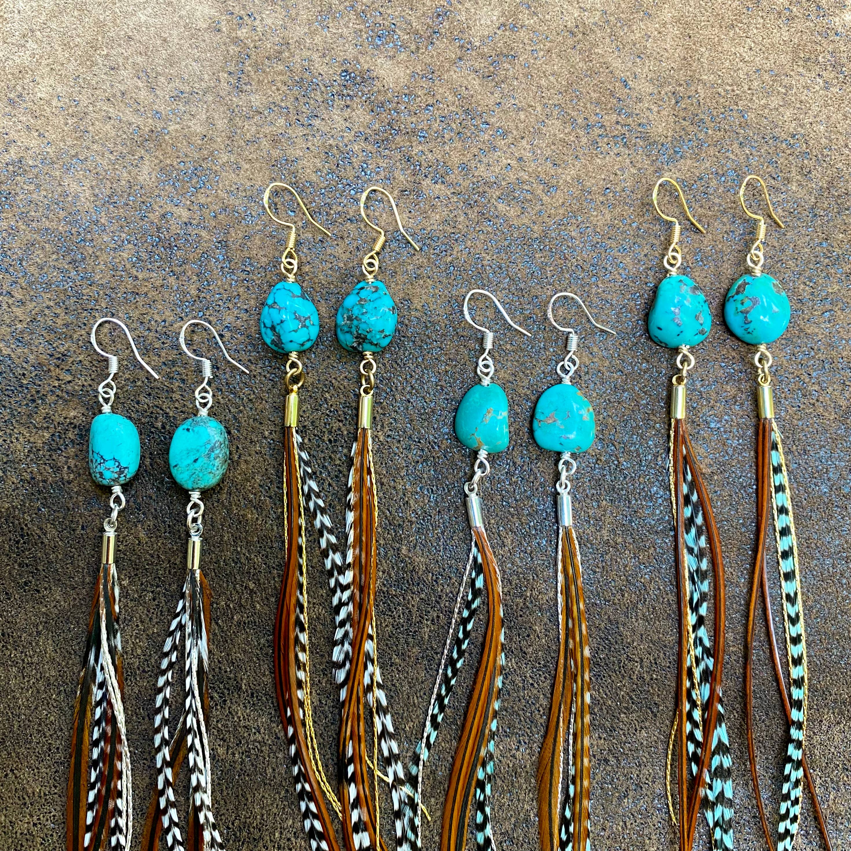 Five pairs of ASTALI Turquoise & Mini Feather Earrings – Grizzly & Fawn/Gold are displayed on a textured brown surface, each featuring turquoise stones, long multicolored feathers, and stylish gold-plated or silver hooks.