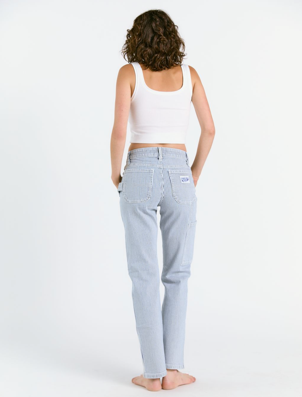 A person with curly hair, wearing a white tank top and Porter Blue Apparels PEYTON PAINTER IN PACIFIC WASH carpenter pant, stands barefoot facing away against a plain white background.