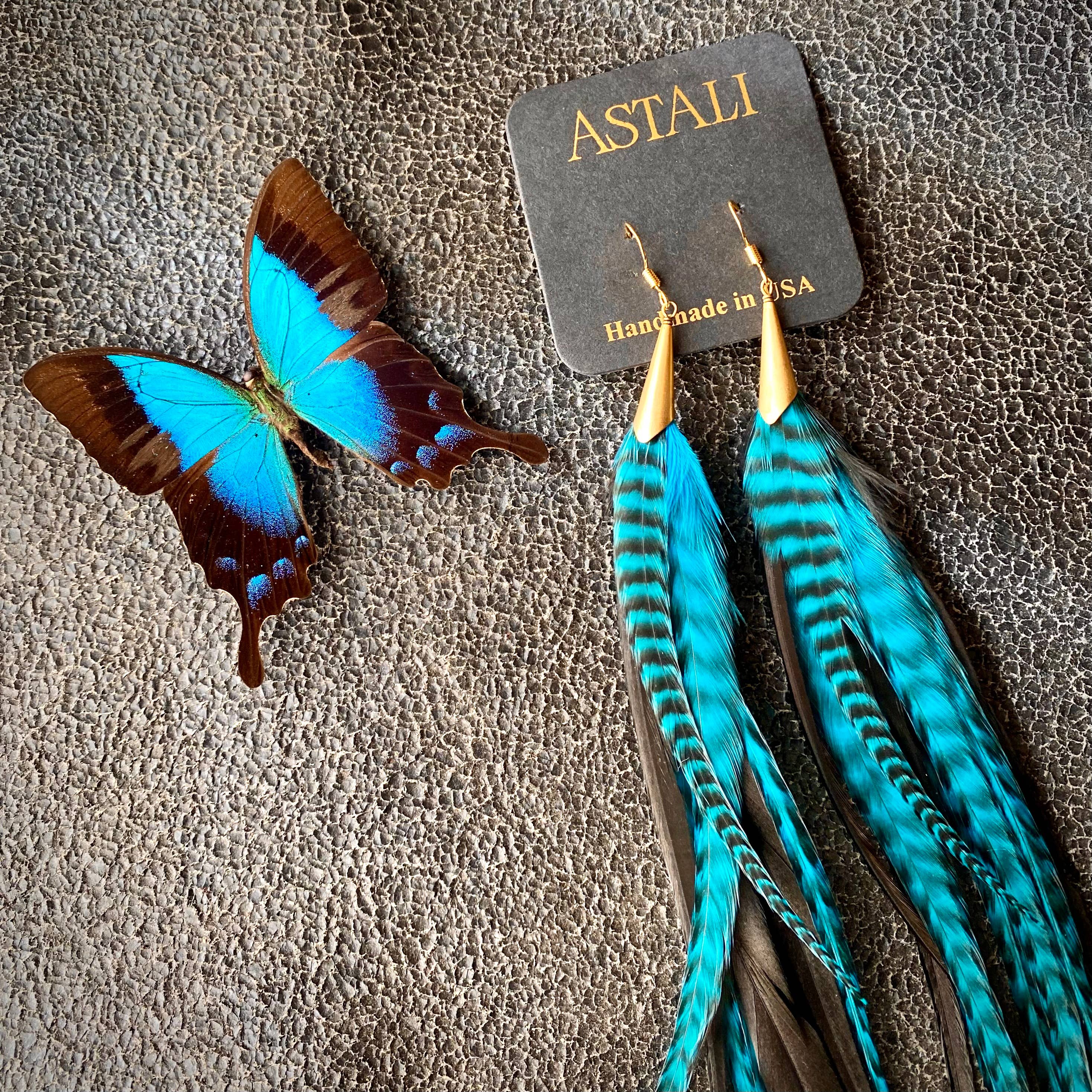 A blue and black butterfly rests beside ASTALI’s Long Feather & Cone Earrings—Turquoise Grizzly & Black/Gold—made from sustainably sourced feathers, displayed on a card labeled ASTALI Handmade in USA.