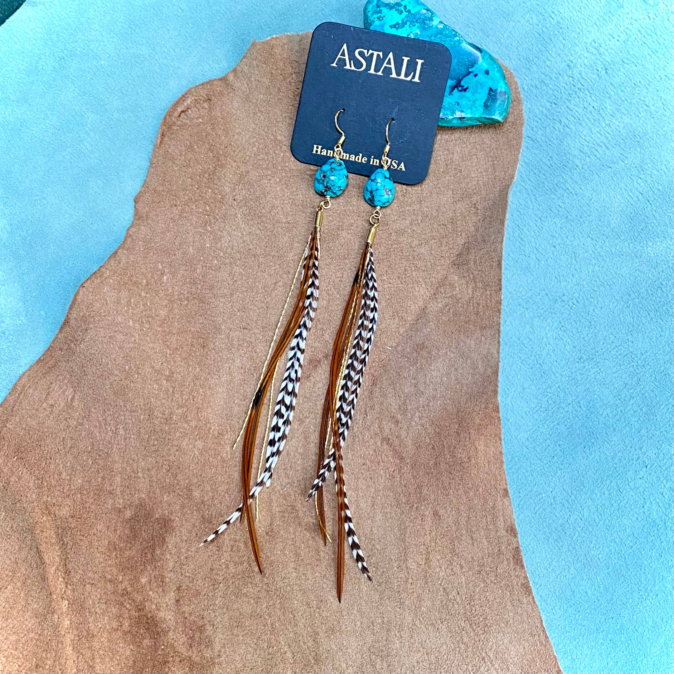 Turquoise & Mini Feather Earrings - Grizzly & Fawn/Gold by ASTALI, handcrafted in the USA, feature turquoise stones and feather accents, displayed on a branded card against a soft brown and blue background.