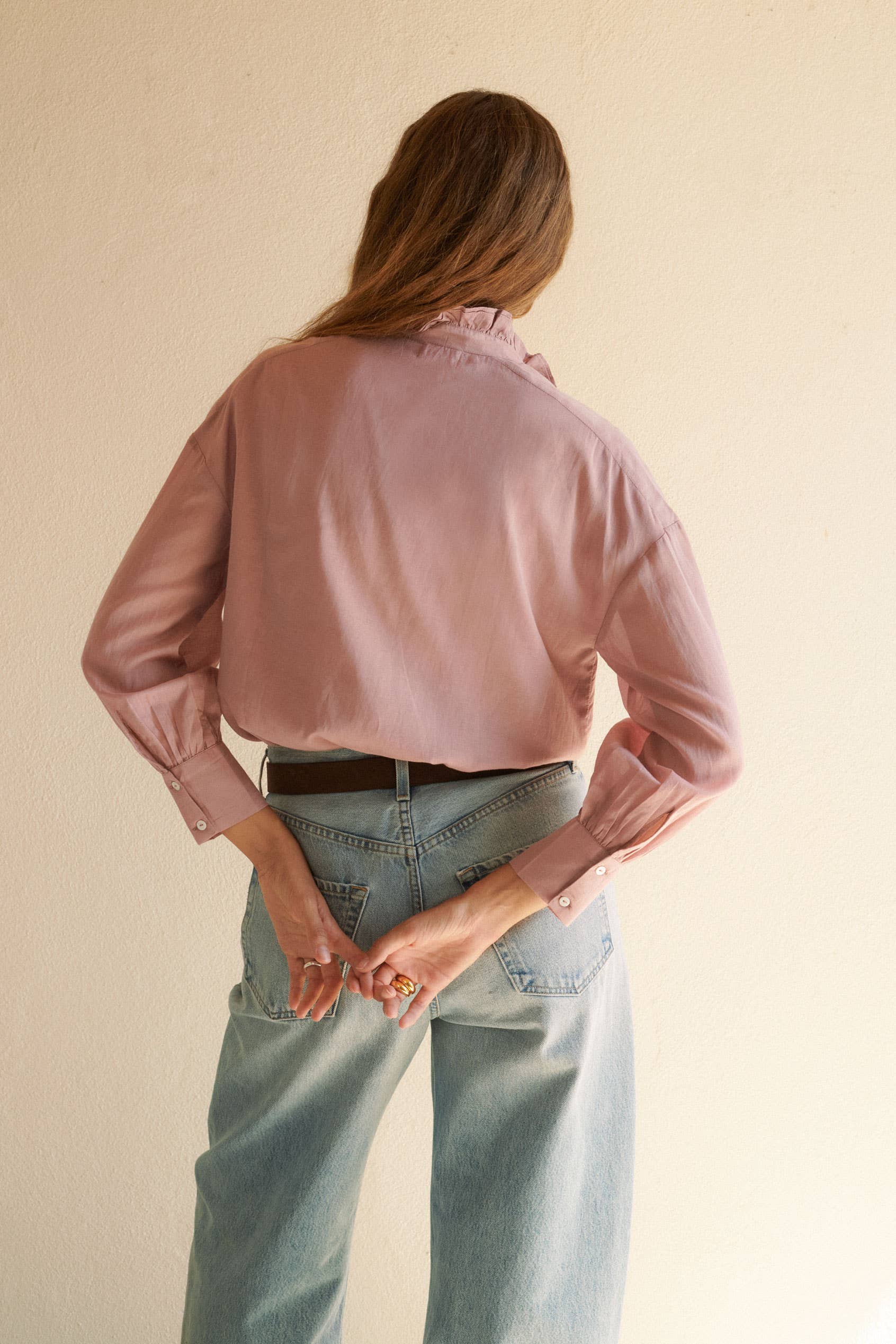 A person stands facing away, wearing the Alexa Blouse Rose by Sonmer Shop USA and light blue jeans. Their hands are clasped behind their back, long brown hair drapes over their shoulders, and the background is a plain beige wall.