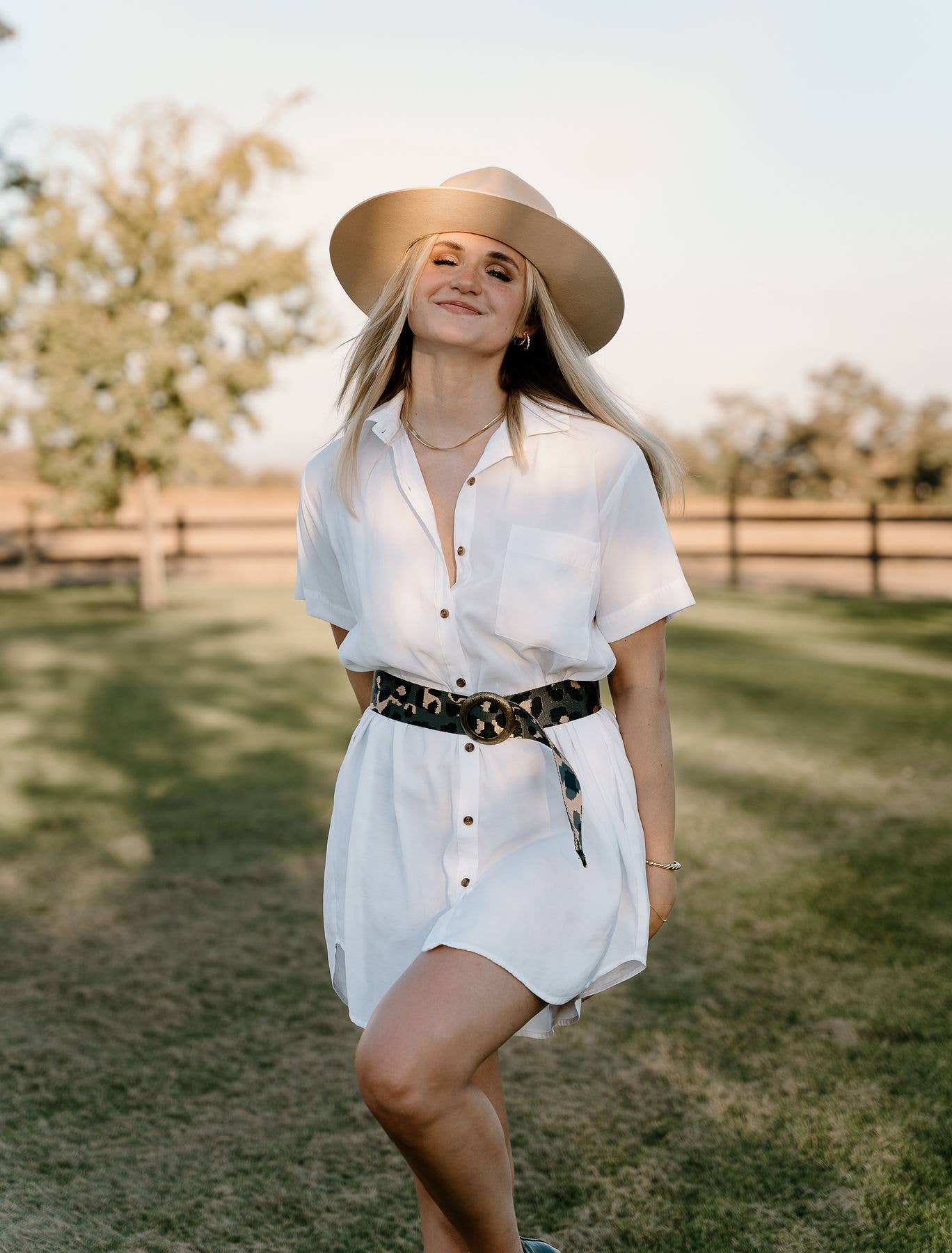 A woman smiles on a grassy lawn, wearing a wide-brimmed hat and a white button-down dress styled with the Ginger Blue Wild Child Leopard belt. Trees and a wooden fence are visible in the background.